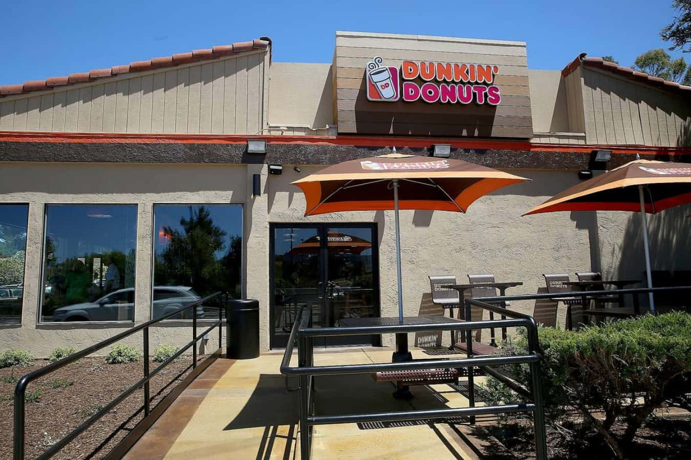 Dunkin Donuts 桌布 壁紙 — Dunkin' Donuts 店鋪前一個晴朗的日子，橙色遮陽傘為戶外座位提供陰涼，上方是清澈的藍天。