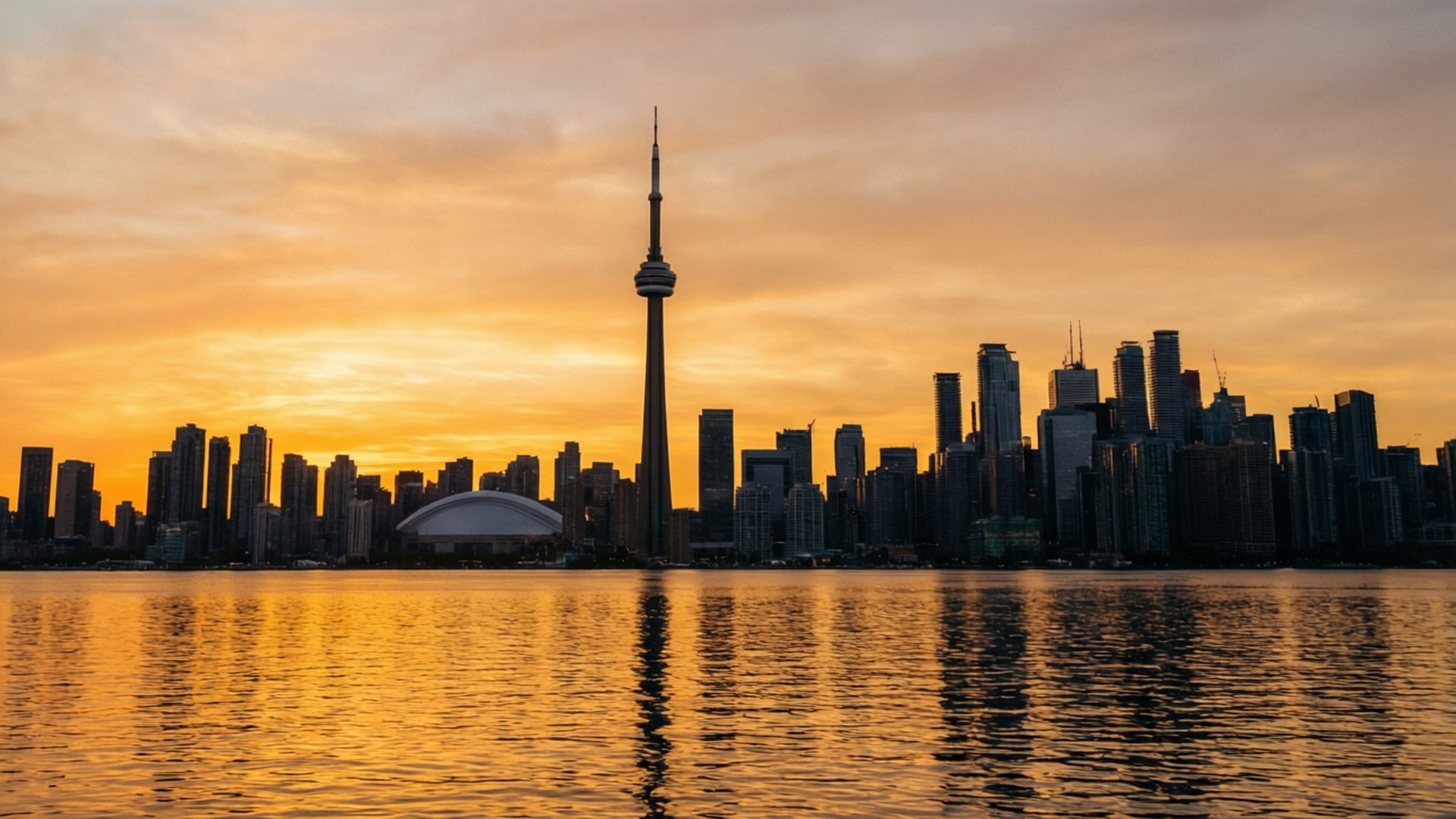Toronto Sunset Skyline 壁紙 — 多倫多天際線在日落時分，CN塔和摩天大樓在金色橙色雲彩的襯托下形成剪影，倒映在平靜的水面上。