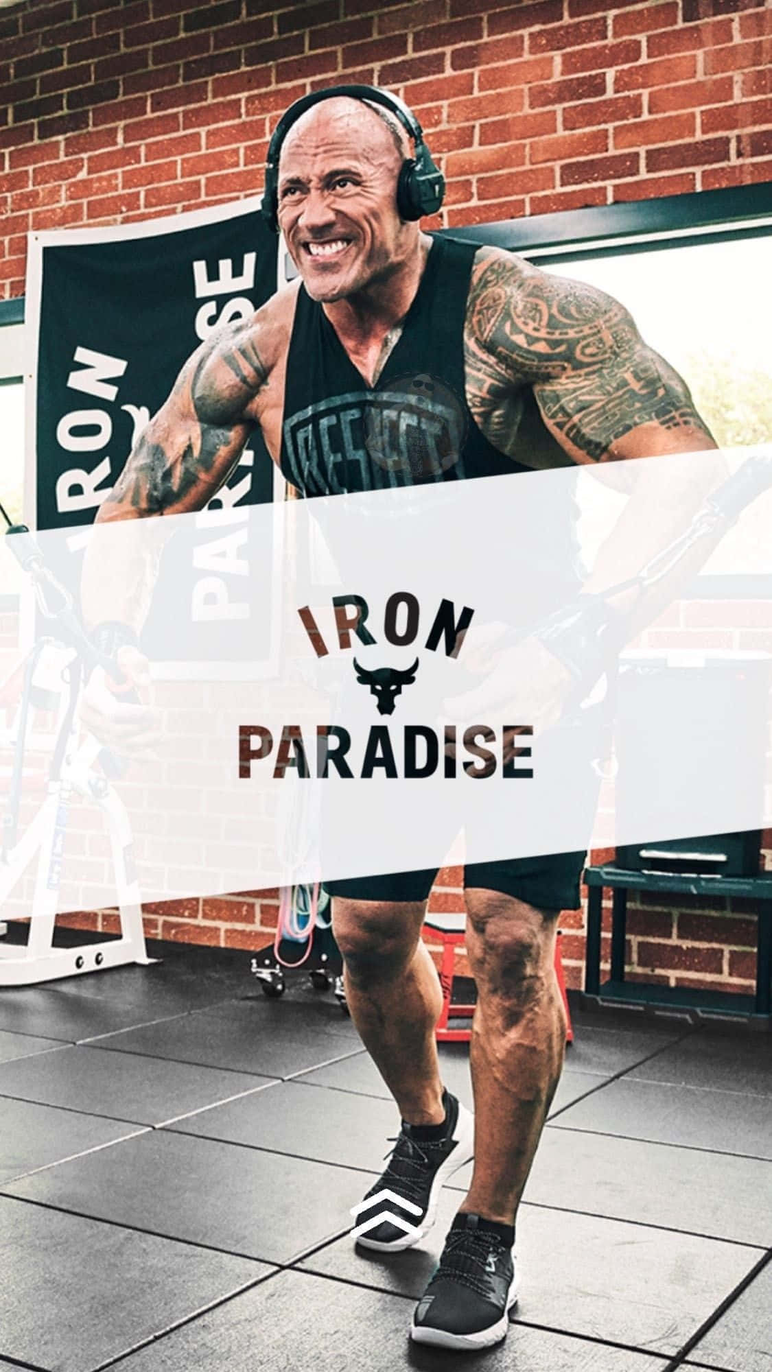 Project Rock 桌布 壁紙 — Dwayne Johnson 在有磚牆和 'Iron Paradise' 品牌的健身房裡舉重時咧嘴笑。