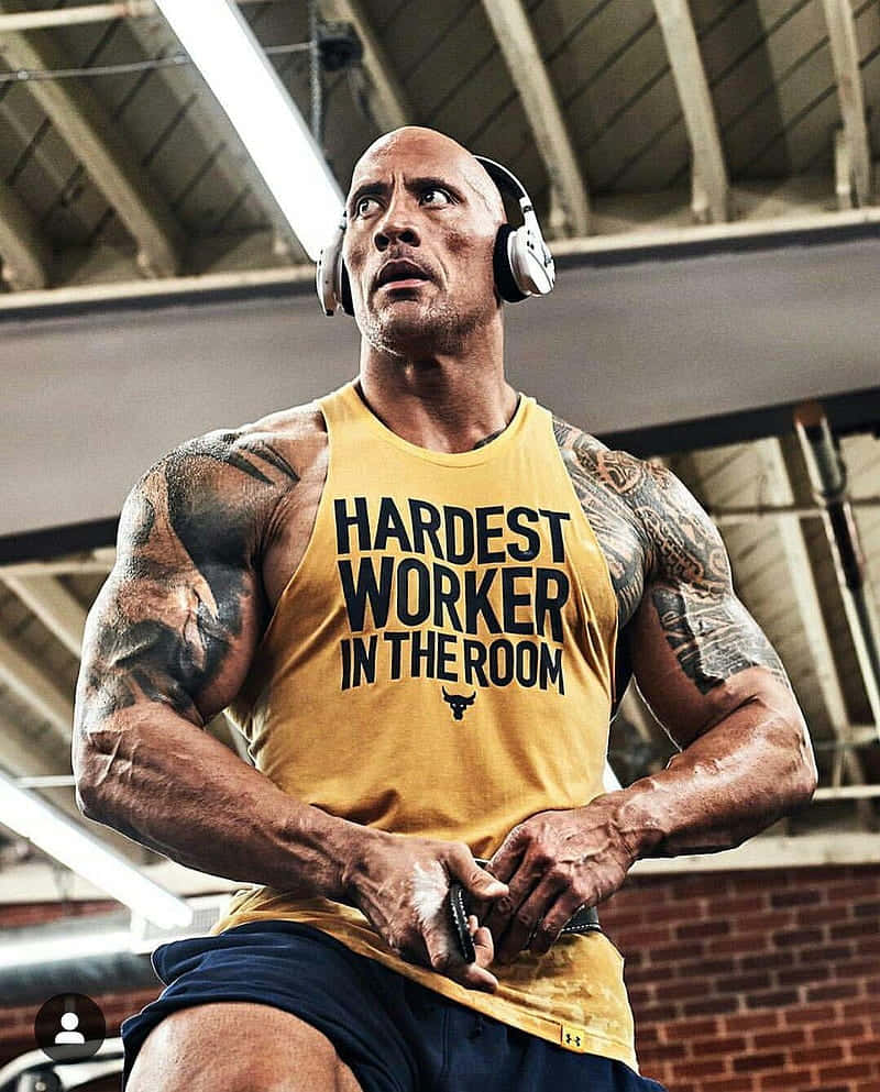 Project Rock 桌布 壁紙 — Dwayne Johnson 在健身房戴著耳機，身穿黃色背心，上面寫著 'hardest worker in the room'。