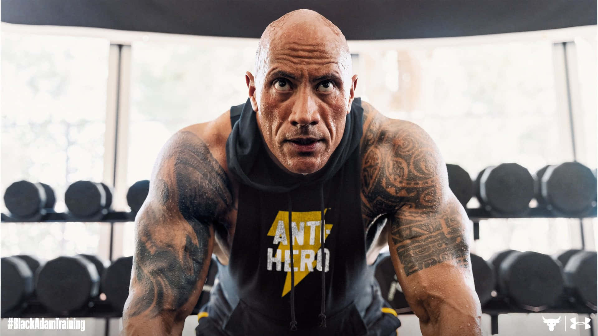 The Rock 桌布 壁紙 — Dwayne Johnson 在健身房穿著帶有黃色閃電和文字的黑色連帽衫，周圍環繞著啞鈴。