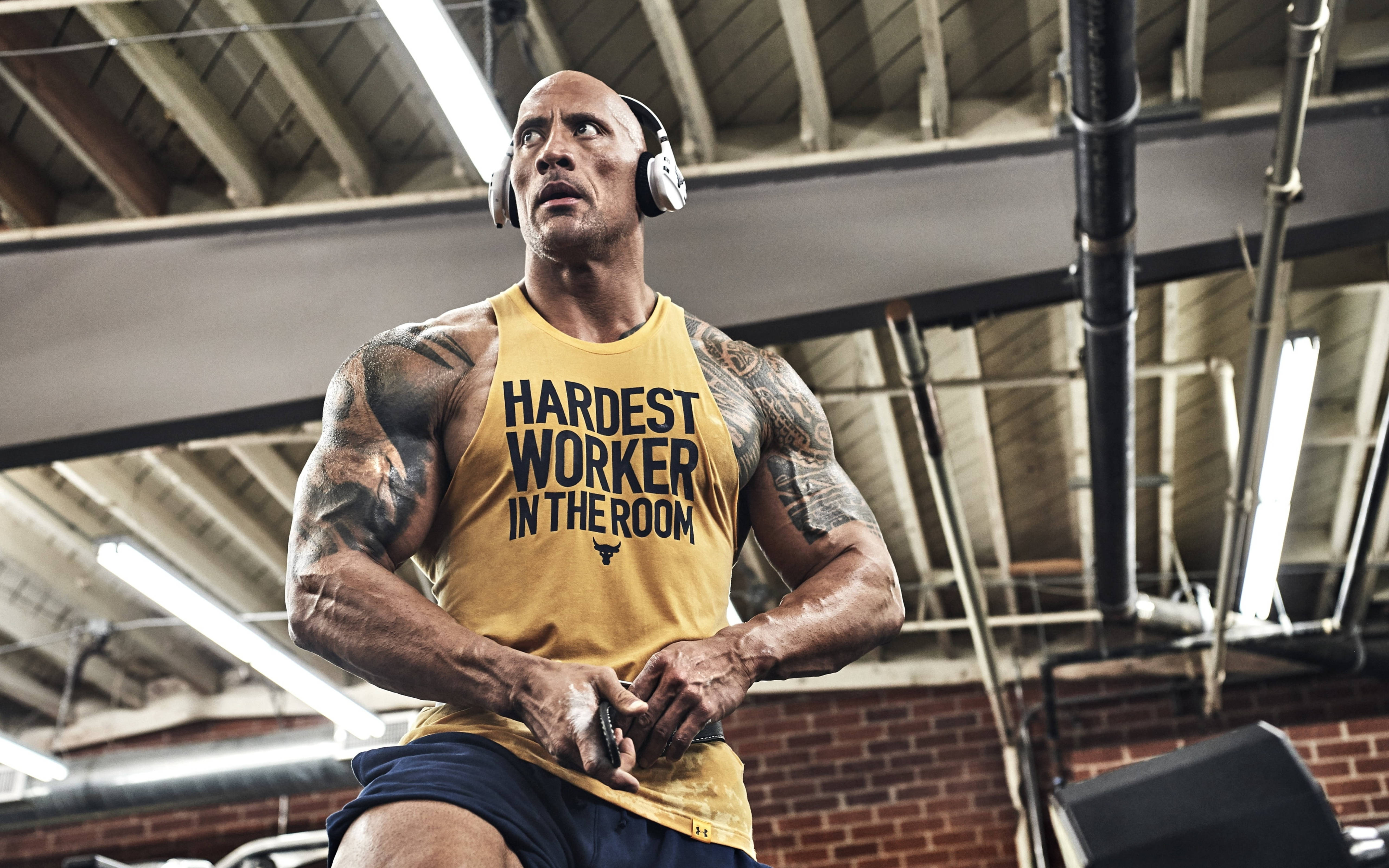 Dwayne 桌布 壁紙 — Dwayne Johnson 在健身房戴著耳機，身穿印有 'Hardest Worker in the Room' 字樣的黃色背心。