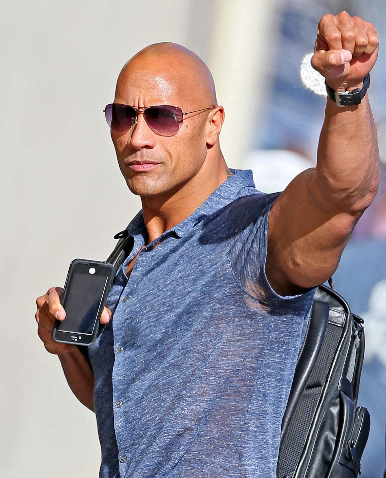 演員 桌布 壁紙 — Dwayne Johnson 戴著太陽眼鏡和藍色襯衫，在戶外自信地拿著電話並舉起拳頭。