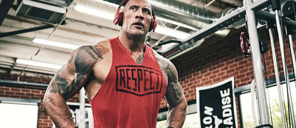 Project Rock 桌布 壁紙 — Dwayne Johnson 身穿紅色 'RESPECT' 背心及耳機，在設有裸露磚牆和器材的健身房中強勢站立。