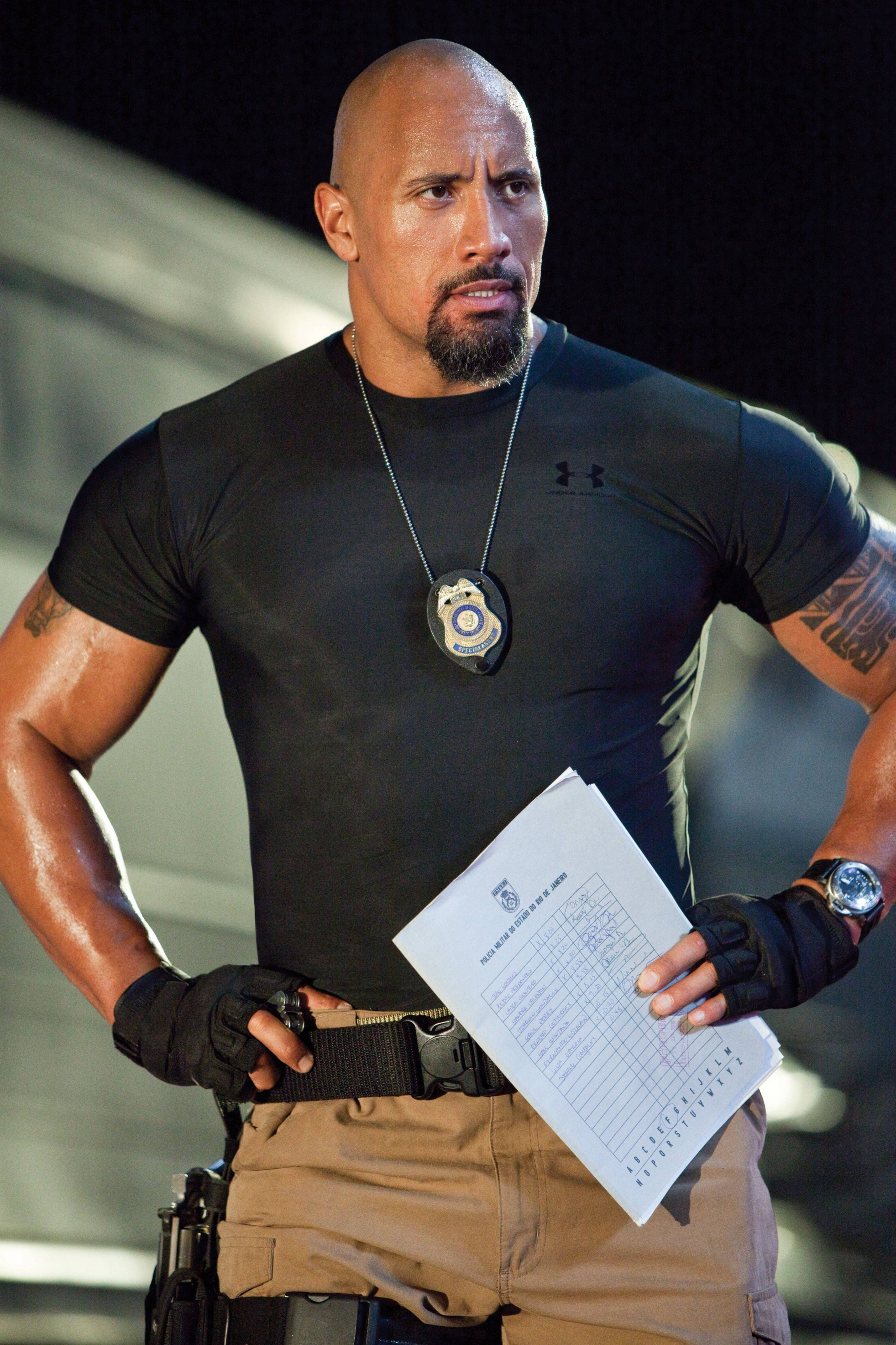 Dwayne 桌布 壁紙 — Dwayne Johnson 飾演一名肌肉發達的警察，身穿黑色襯衫，手持報告，表情嚴肅。