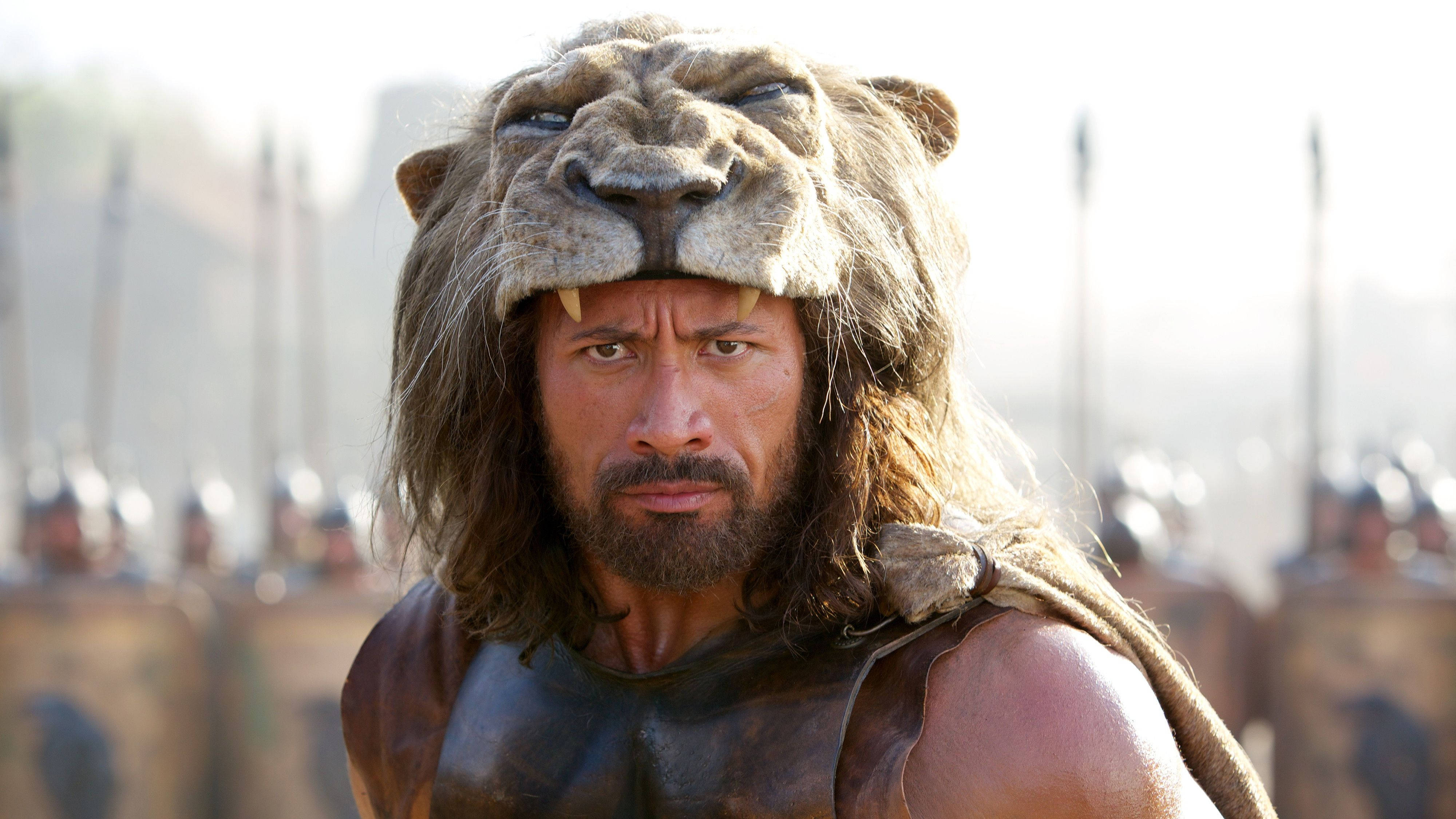 海克力斯 桌布 壁紙 — Dwayne Johnson 飾演的戰士戴著獅子頭盔，挑釁地站在模糊的士兵軍隊前面。