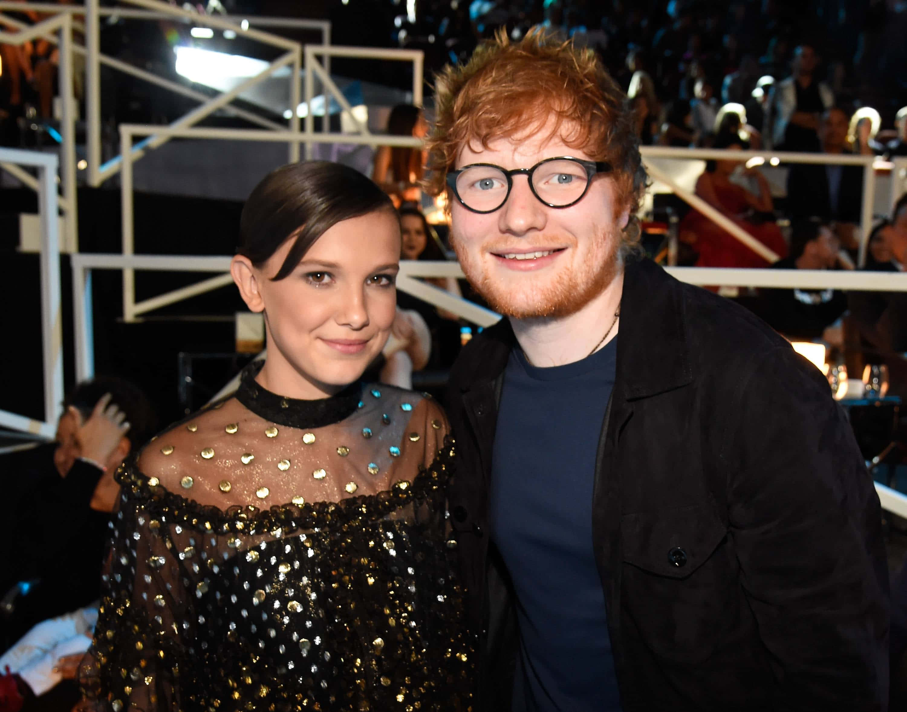 米莉·芭比·布朗 桌布 壁紙 — Ed Sheeran 和 Millie Bobby Brown 在明亮的舞台燈光下於一場華麗的活動中一起微笑。