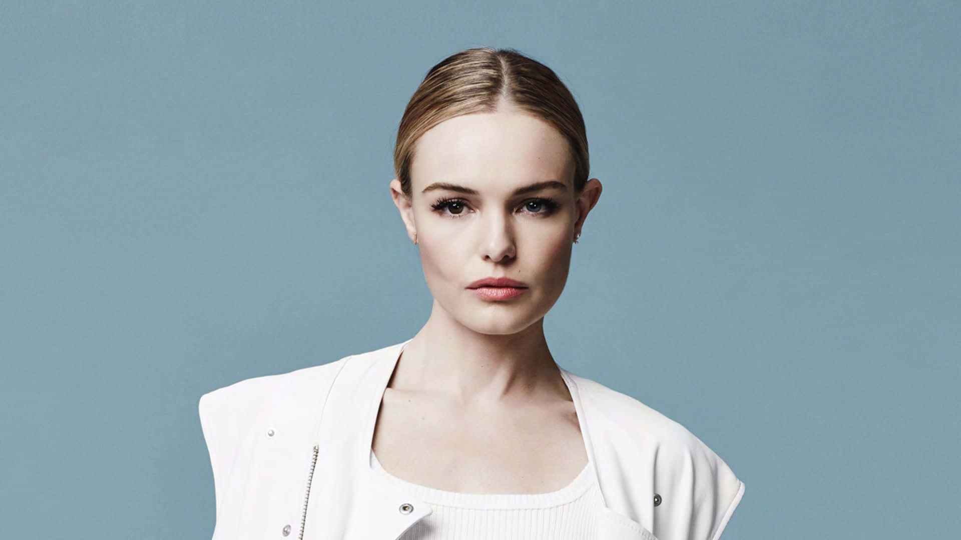 Kate Bosworth 桌布 壁紙 — 一位優雅的金髮女性，頭髮向後梳，穿著白色外套，背景是純藍色攝影棚。
