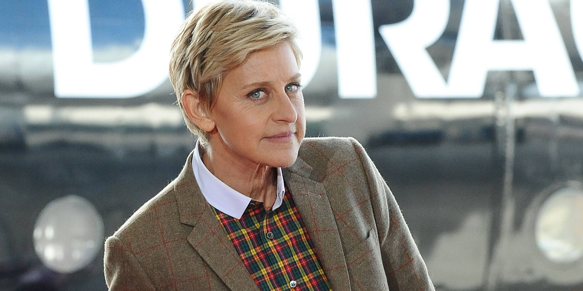 艾倫·狄珍妮 桌布 壁紙 — Ellen DeGeneres 身穿棕色粗花呢外套和格紋襯衫，站在一輛帶有「DURACELL」標誌的模糊復古汽車前。