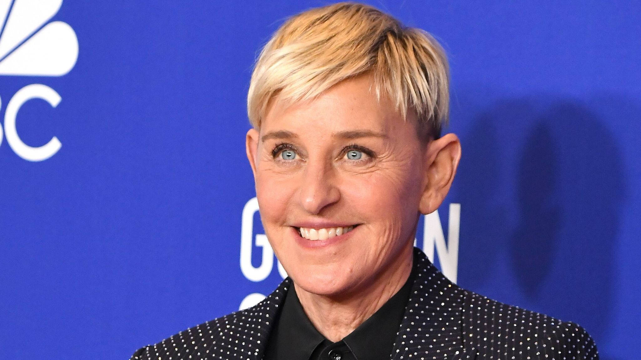 Ellen Degeneres 桌布 壁紙 — Ellen DeGeneres 在活動中穿著黑色圓點西裝外套，在藍色NBC背景下溫暖地微笑。