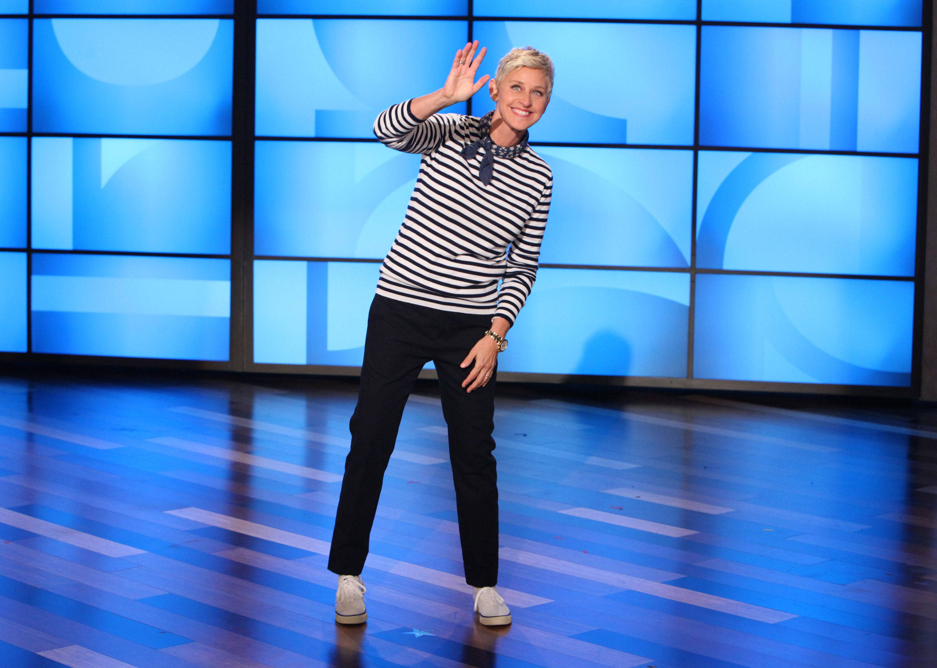 Celebrity 桌布 壁紙 — Ellen DeGeneres 在藍色燈光的攝影棚舞台上，以幾何螢幕背景愉快地揮手。