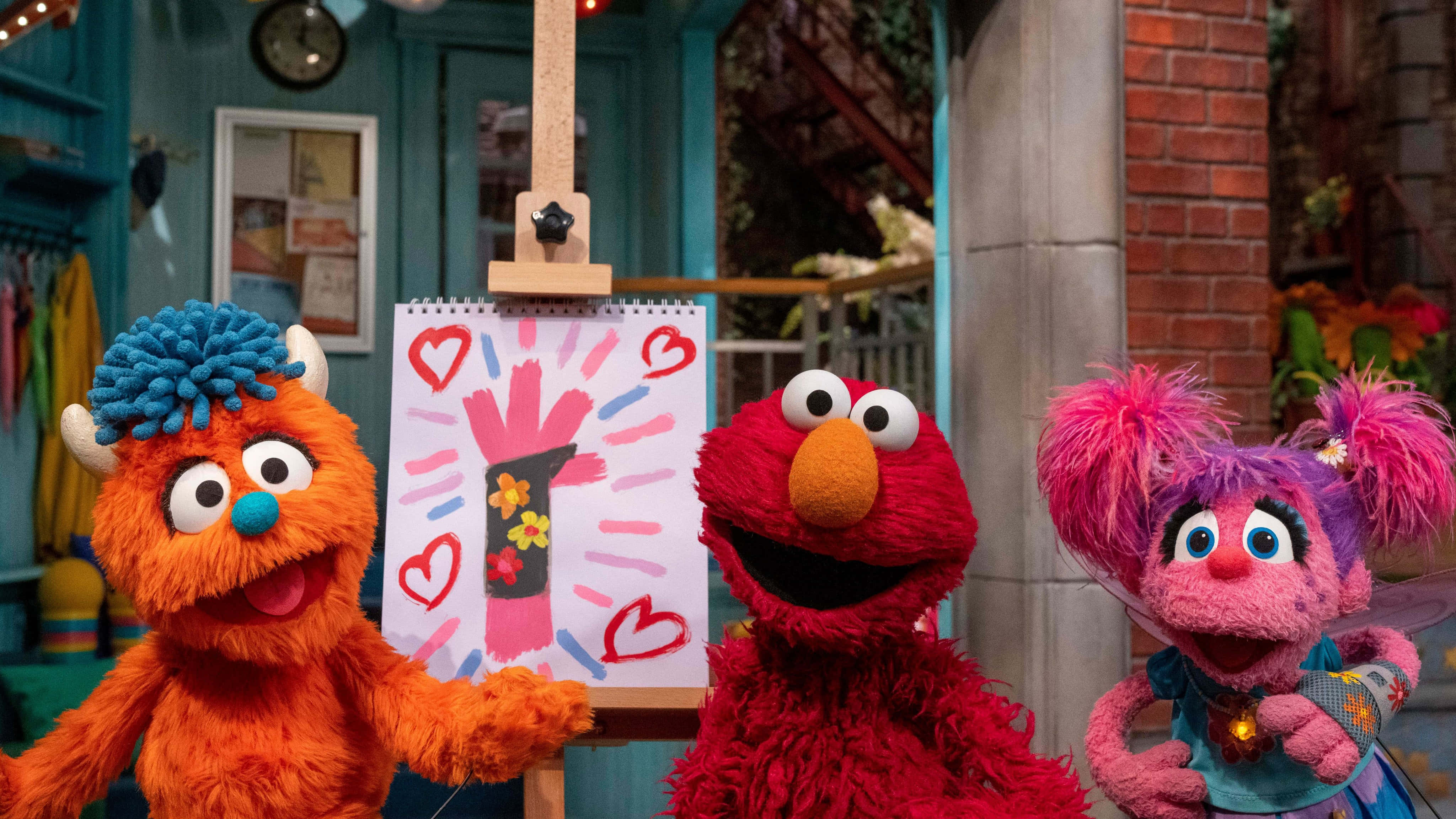 Elmo 桌布 壁紙 — Elmo、Abby 和一隻毛茸茸的怪物在色彩繽紛的場景中，愉快地在畫架上的童趣畫作旁擺姿勢。