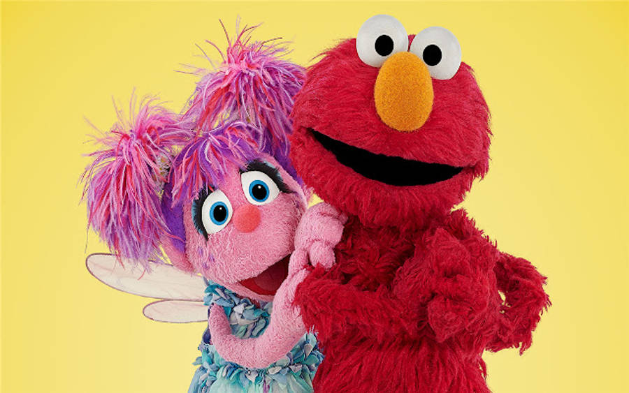 Sesame Street 桌布 壁紙 — Elmo 和一個有紫色辮子的粉紅色仙女玩偶在純黃色背景前溫暖擁抱。