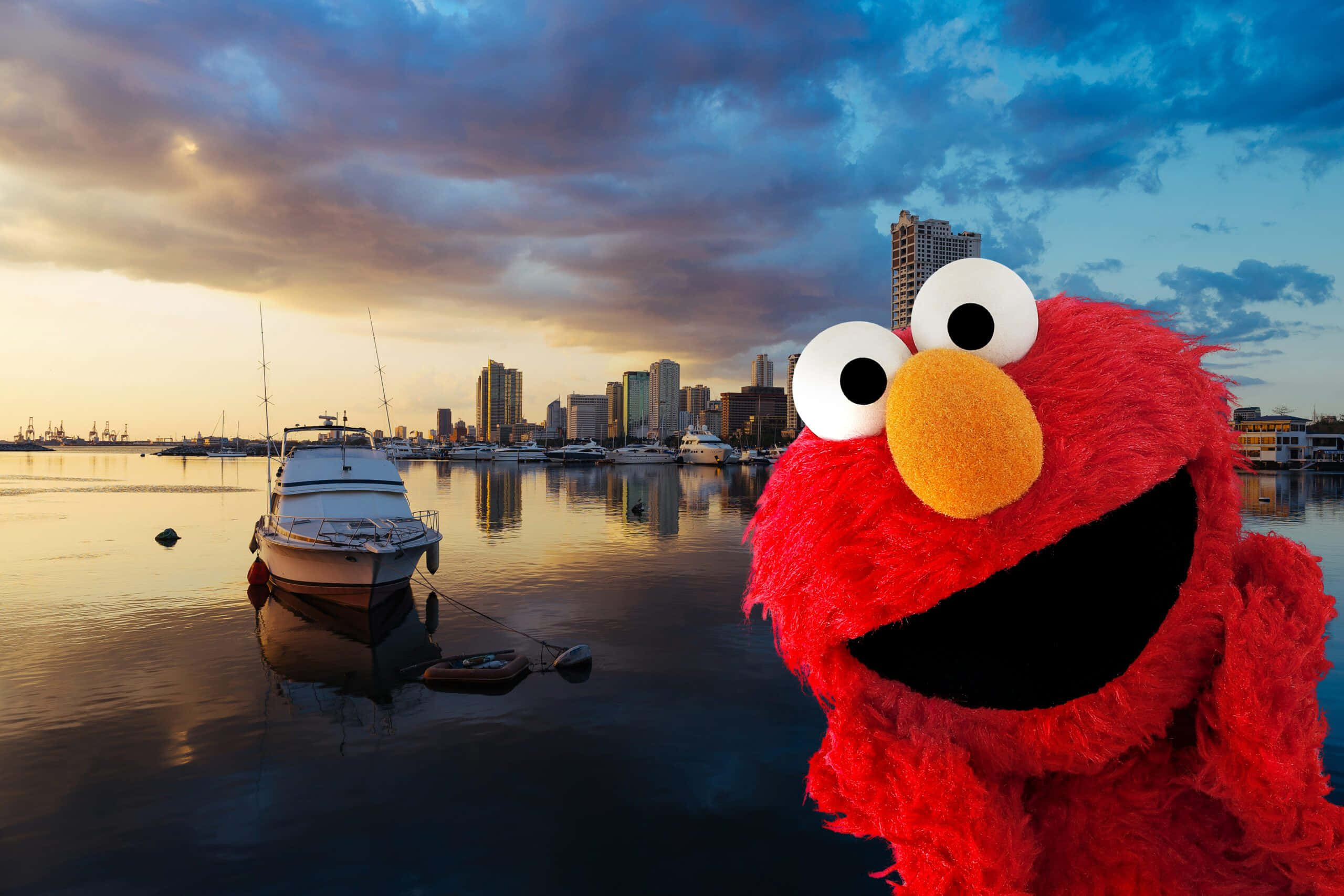 Elmo 桌布 壁紙 — Elmo 在日落時分的港口前燦爛微笑，船隻和城市天際線倒映在平靜的水面上。