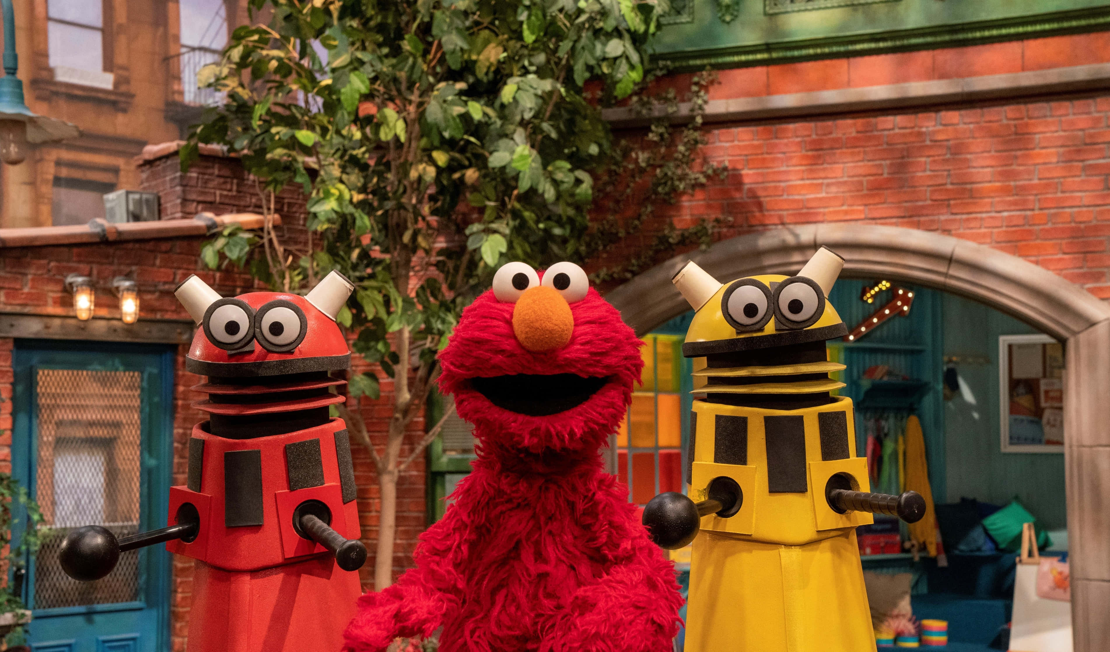 Elmo 桌布 壁紙 — Elmo 愉快地站在兩個色彩繽紛的機器人玩偶之間，背景是充滿活力的芝麻街庭院，有磚造建築和綠植。
