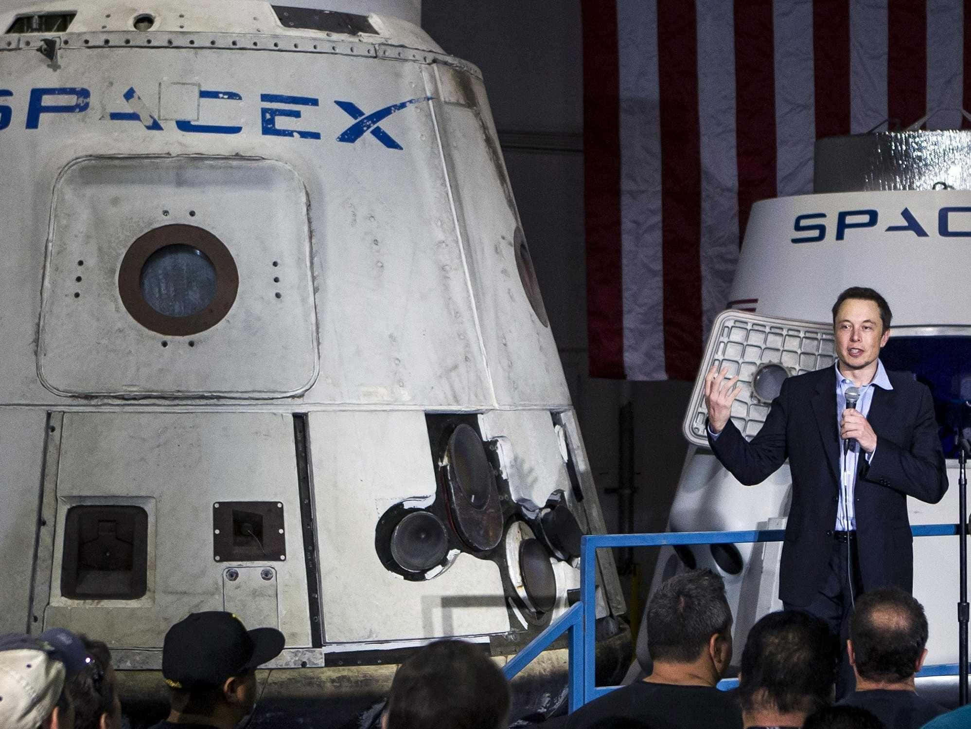 伊隆·馬斯克 桌布 壁紙 — Elon Musk 在室內活動中，身後有美國國旗，在SpaceX太空船旁做手勢。