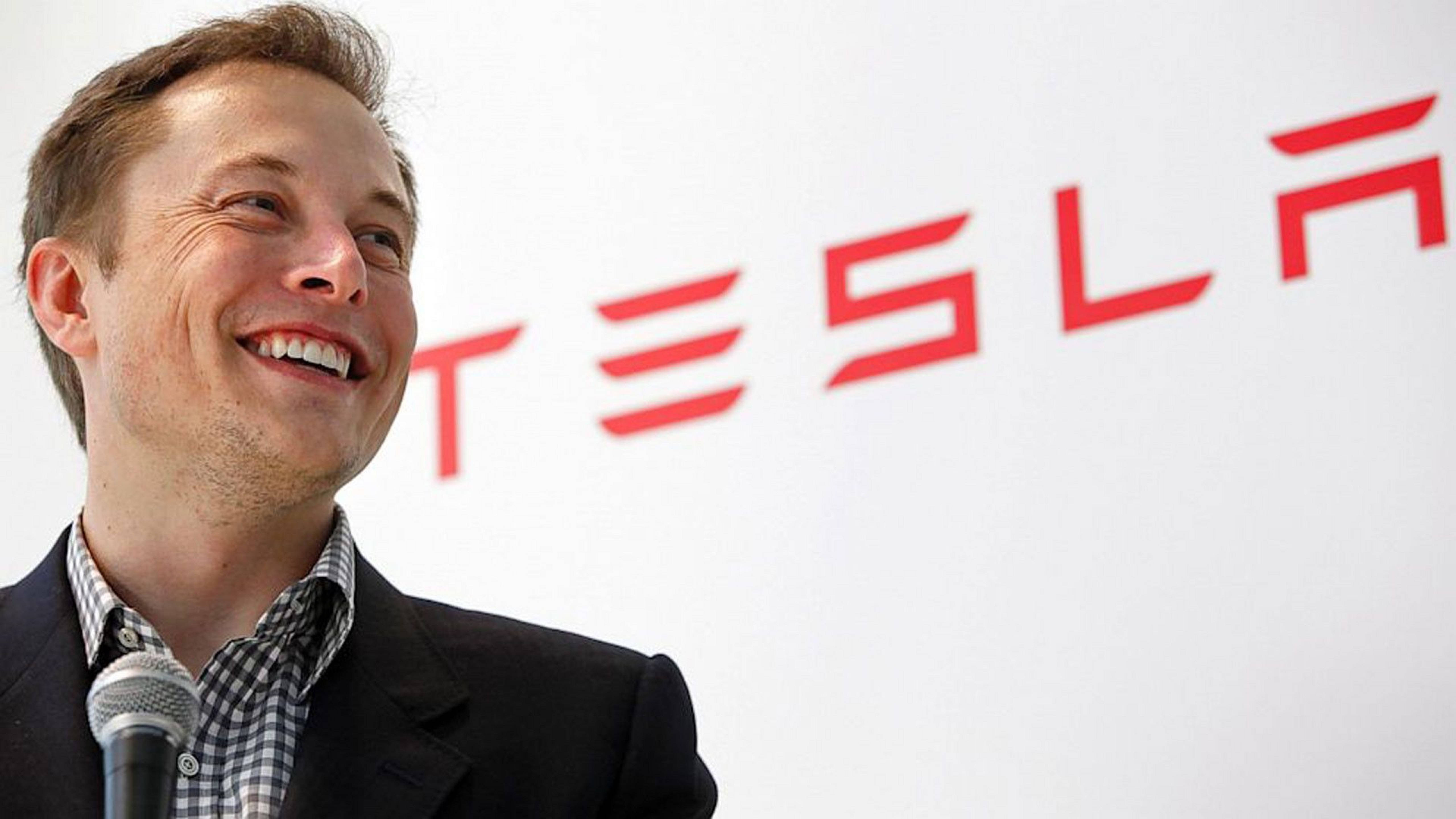 Elon 桌布 壁紙 — Elon Musk 在白色牆壁上的大型紅色 Tesla 標誌前，對著麥克風說話時露出燦爛的笑容。