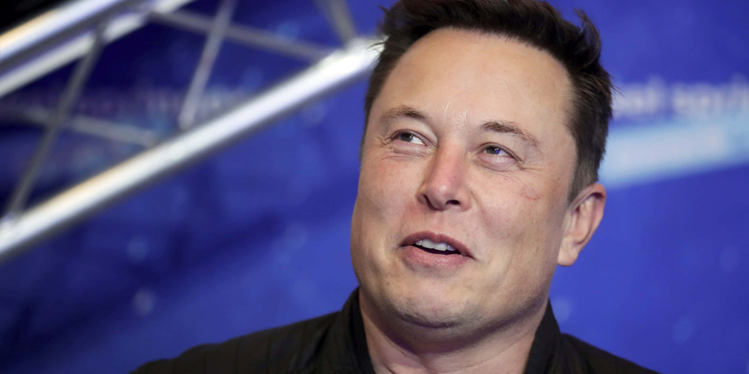 伊隆·馬斯克 桌布 壁紙 — Elon Musk 的特寫鏡頭，向上仰望，表情若有所思，背景為藍色及金屬桁架結構。