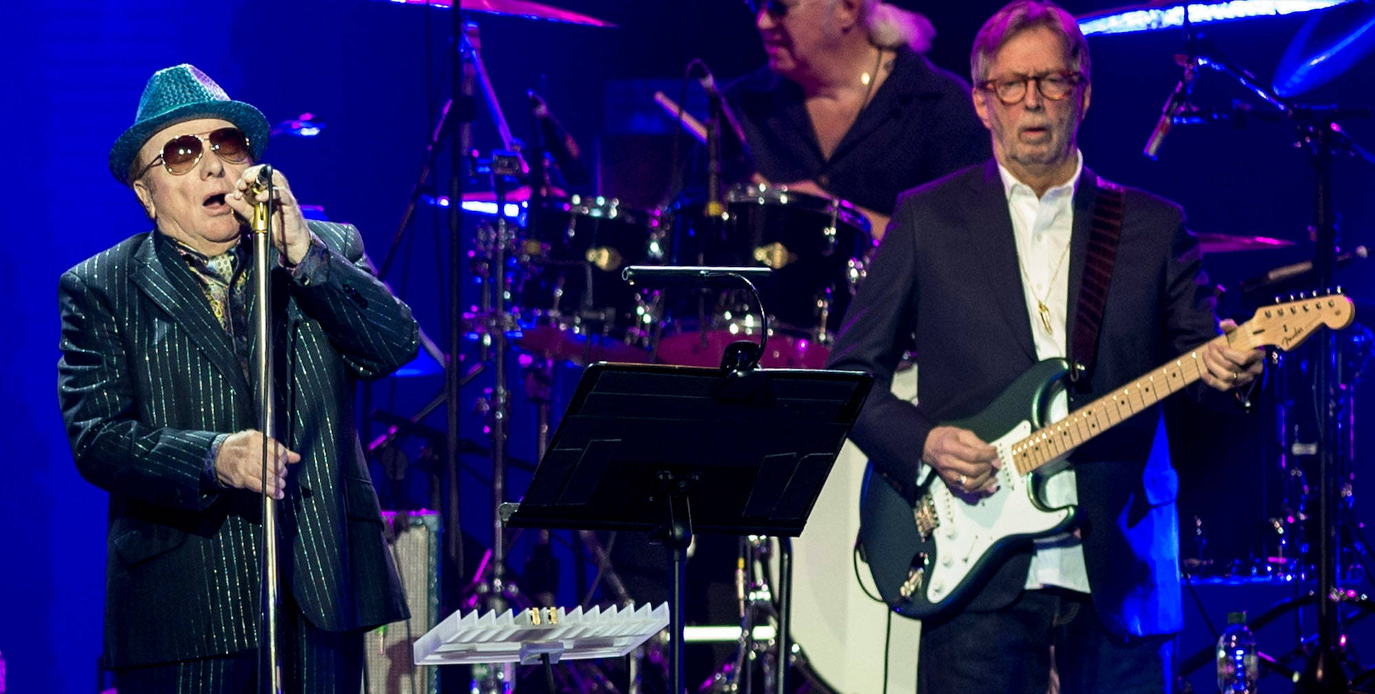 concert 桌布 壁紙 — Elvis Costello 對著麥克風唱歌，吉他手在藍色舞台燈光下演奏，後面有鼓手。
