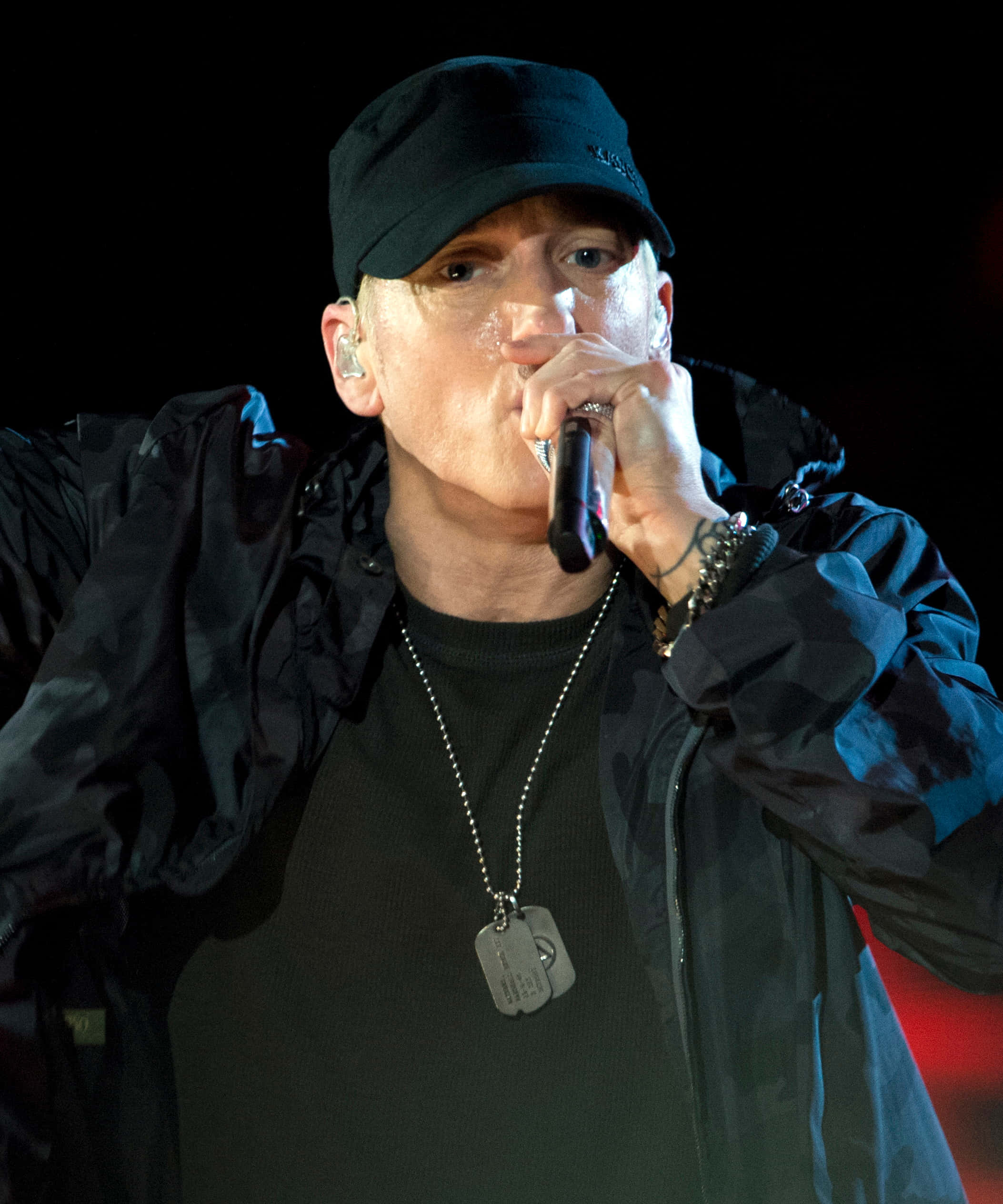 阿姆 桌布 壁紙 — Eminem 在舞台上表演，戴著黑色帽子和外套，在明亮的舞台燈光下將麥克風靠近嘴邊。