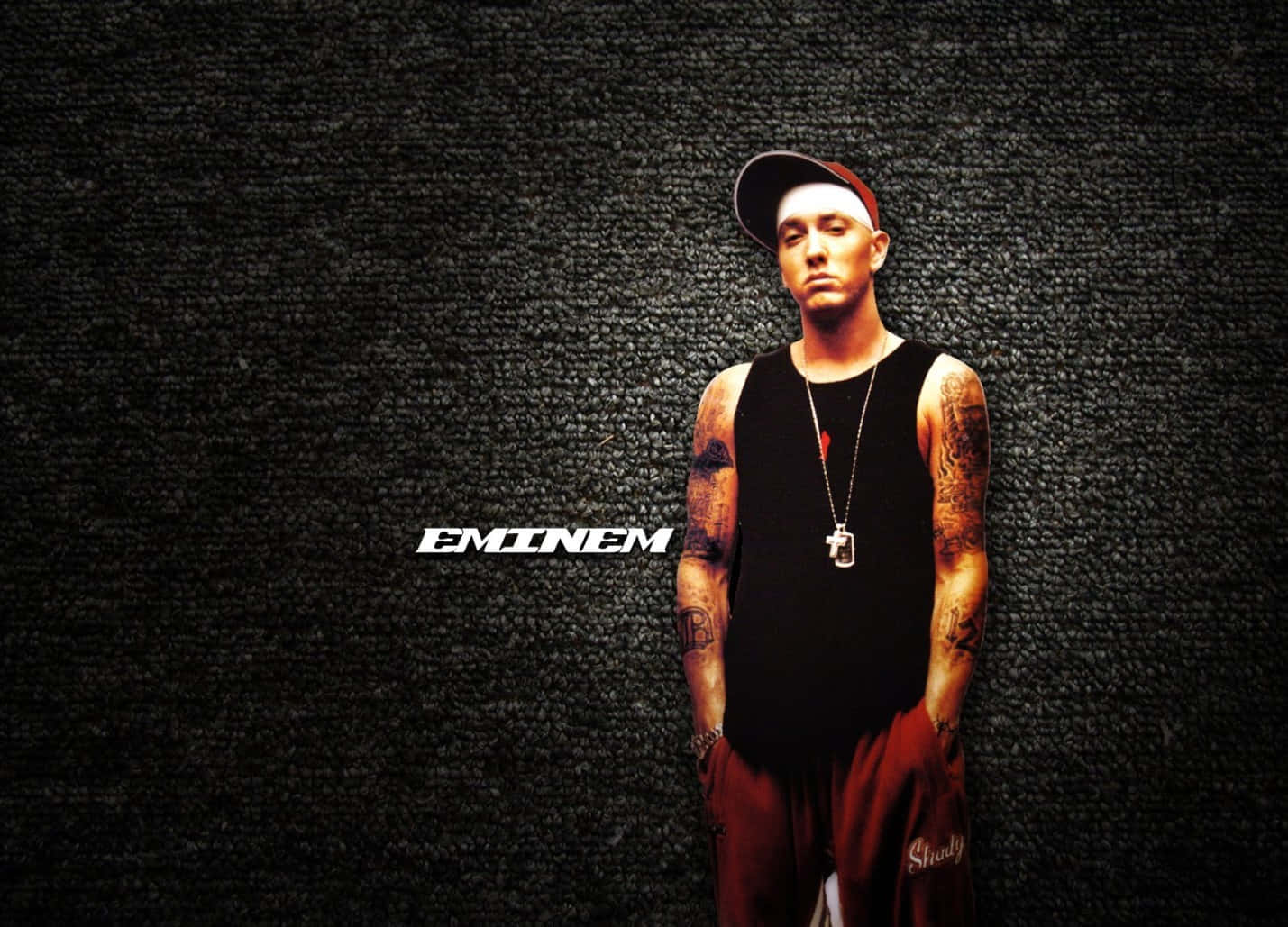 阿姆 桌布 壁紙 — Eminem 自信地站在深色紋理牆前，身穿黑色背心、紅色褲子和帽子，手臂上的刺青清晰可見。