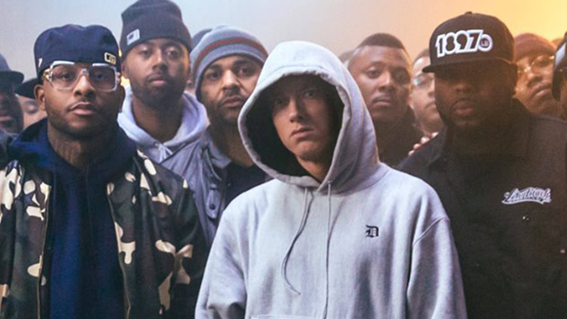 90s Rapper 桌布 壁紙 — Eminem 身穿灰色連帽衫站在中央，被一群穿著都市街頭服飾的嚴肅男子包圍，在溫暖朦朧的燈光下。