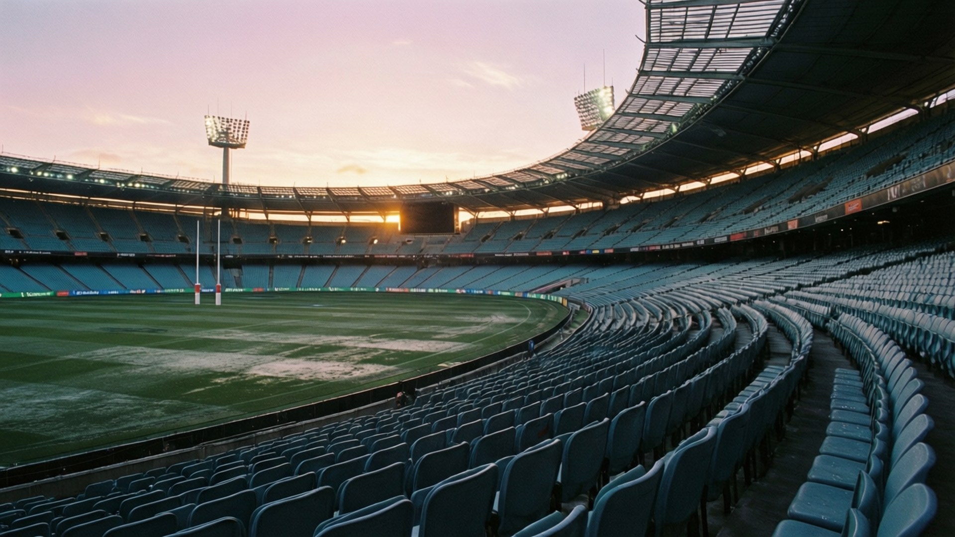 Empty Stadium Dawn 壁紙 — 一個空蕩蕩的體育場在日落時分，藍色座椅排成弧形圍繞著綠色場地，天空呈現粉紅色和橙色。