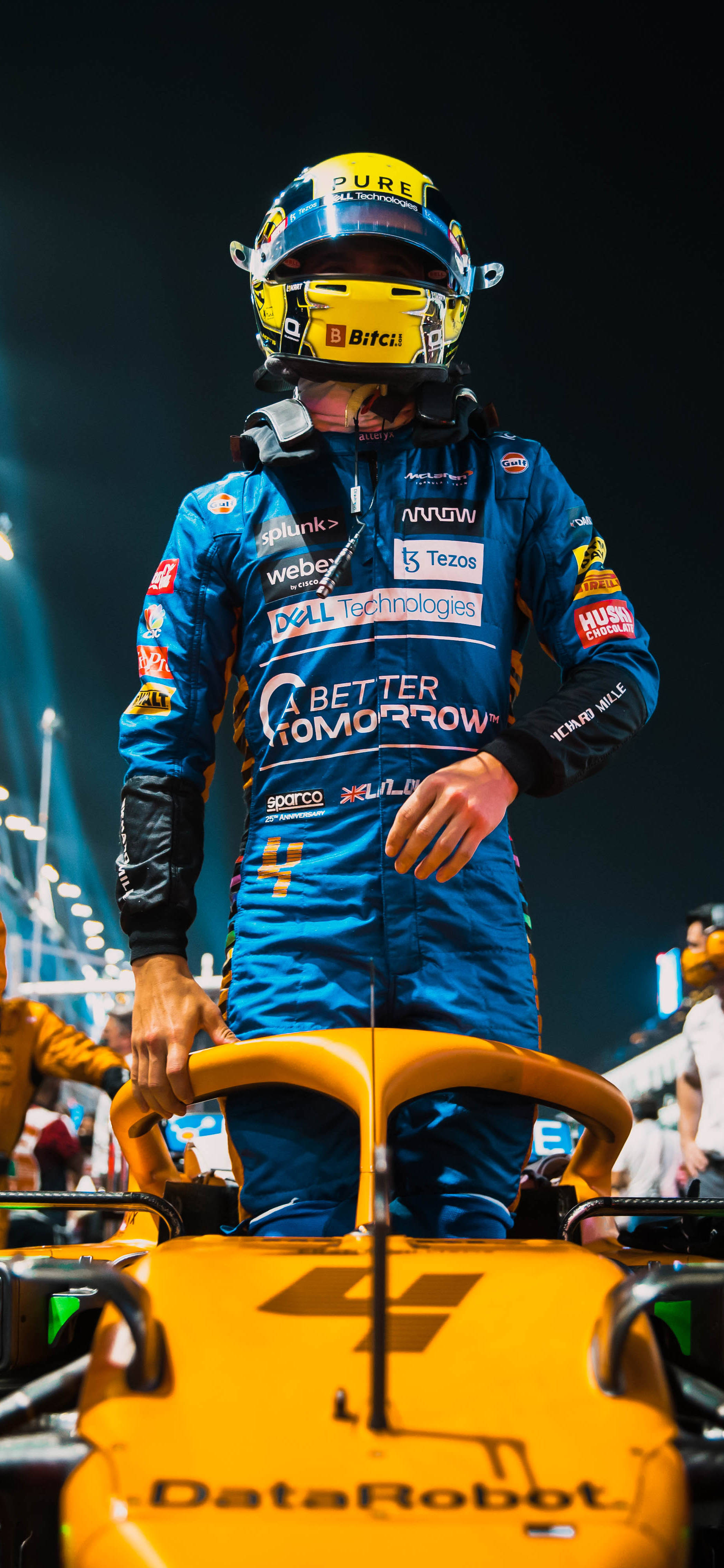 F1 Norris 2021沙烏地阿拉伯GP iPhone 壁紙 — 一位身穿藍色賽車服的F1車手在夜間體育場燈光下站在他亮黃色的賽車上。