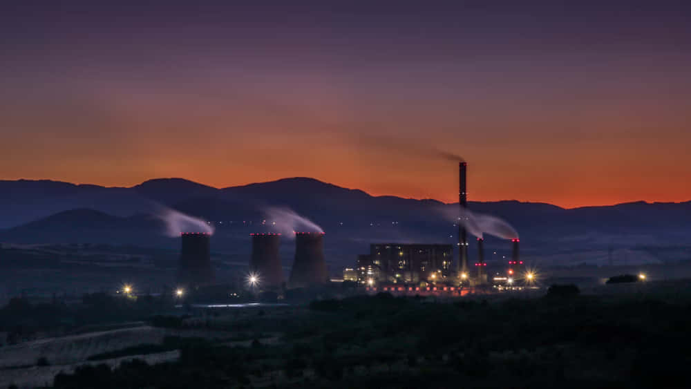  壁紙 — 一座設有冷卻塔的發電廠在遠山之上，向充滿活力的橙色和紫色暮色天空排放蒸汽。