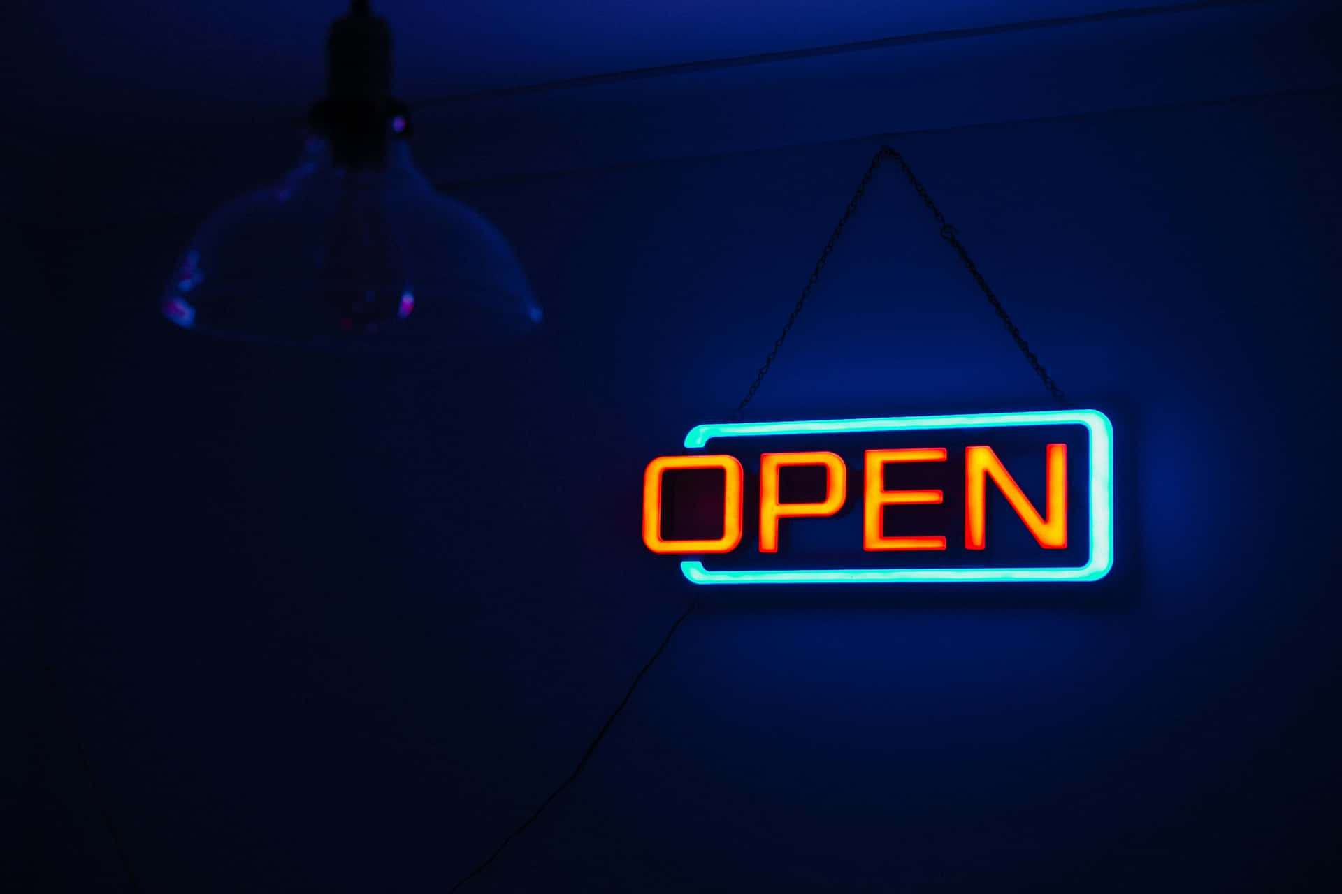  壁紙 — 一個發光的橙色和藍色霓虹燈 'OPEN' 標誌掛在黑暗的牆上，下方有一個復古燈泡。