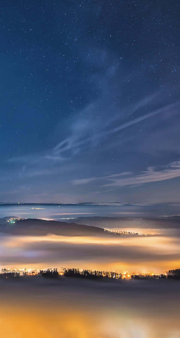  壁紙 — 繁星點點的夜空拱形籠罩著被發光薄霧覆蓋的山谷，遠處城鎮的燈光在下方閃爍。