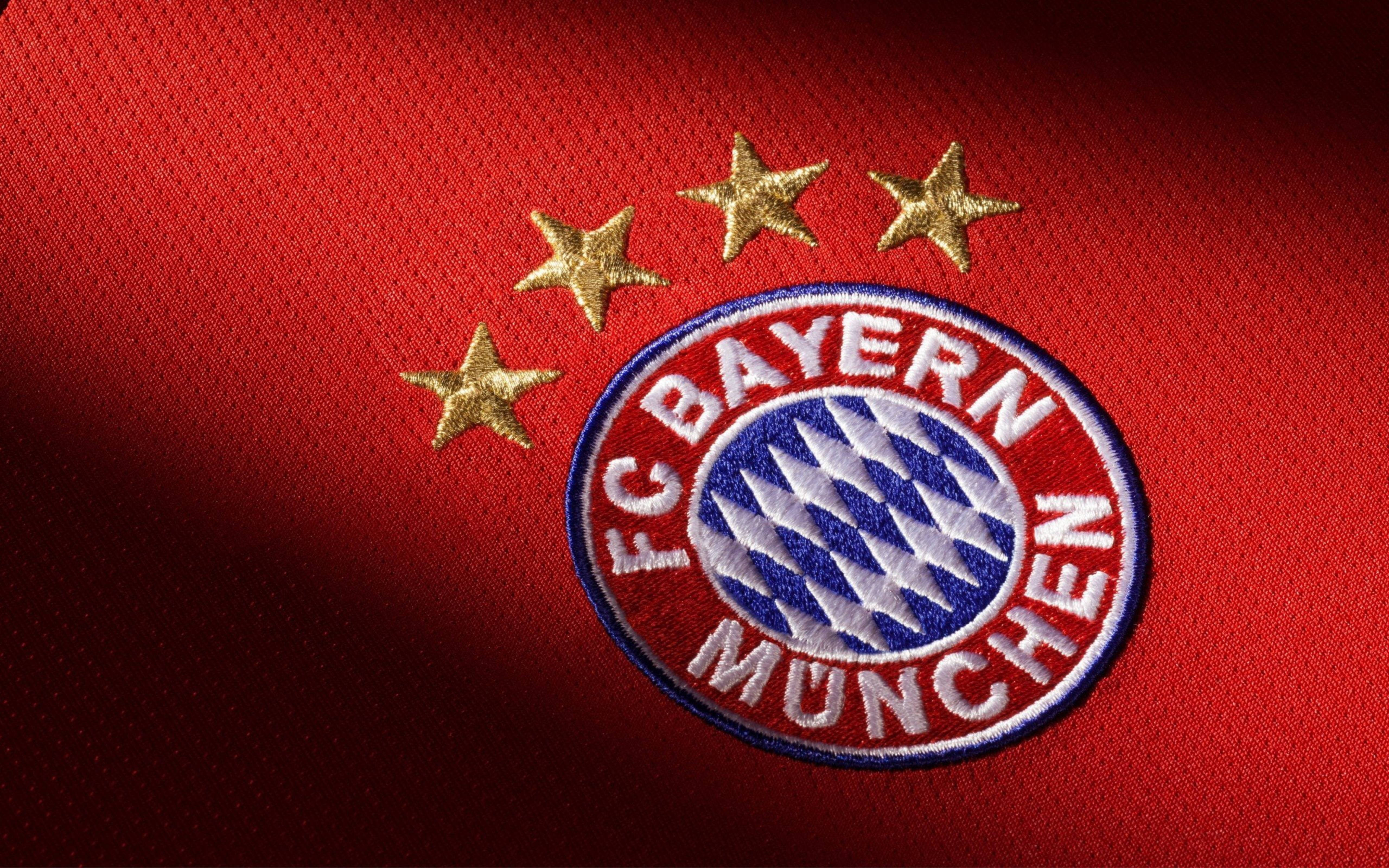 Bayern 桌布 壁紙 — FC拜仁慕尼黑隊徽特寫，四個金色星星刺繡在豐富的紅色布料上，在戲劇性燈光下。
