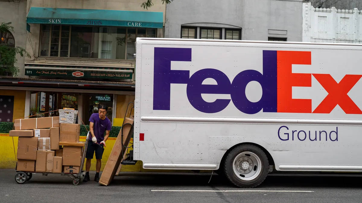 聯邦快遞 桌布 壁紙 — FedEx Ground 司機在城市街道上，將裝滿盒子和一個大型扁平物品的手推車推到他送貨卡車旁邊。
