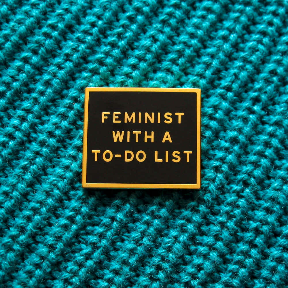 壁紙 — 一個方形黑色琺瑯別針，上面有金色文字寫著「FEMINIST WITH A TO-DO LIST」，放置在帶有紋理的青綠色針織布料上。