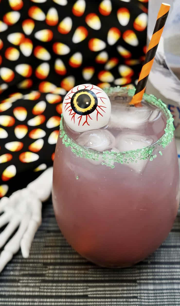 Halloween Drinks 桌布 壁紙 — 一杯粉紅色萬聖節雞尾酒，帶有糖果玉米邊緣和眼球裝飾，旁邊是骷髏手和糖果玉米圖案背景。