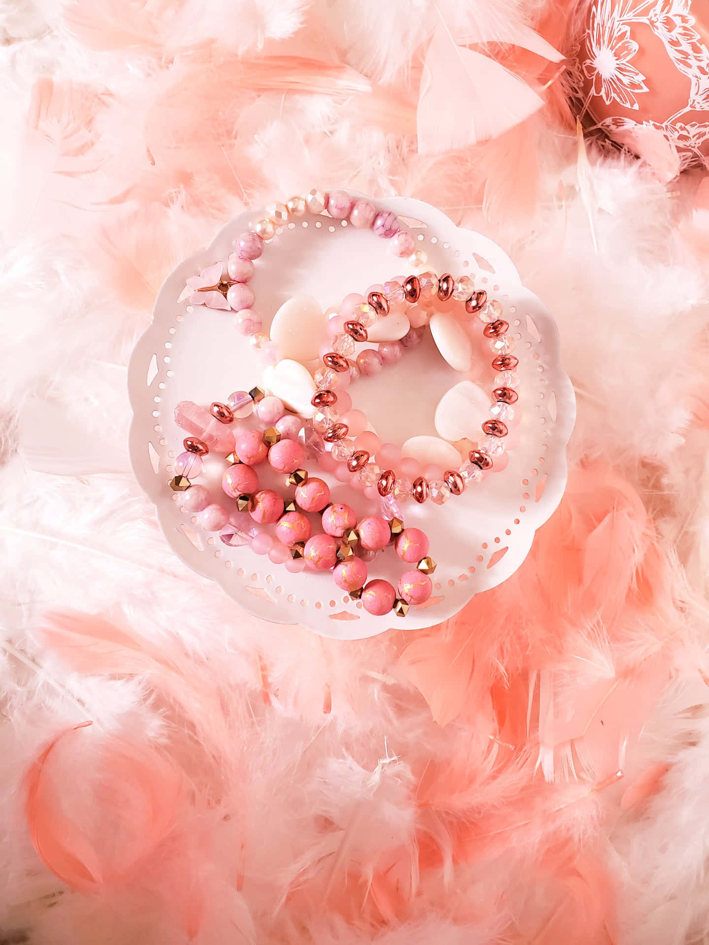  壁紙 — 粉紅色珠子手鐲放在白色扇貝邊碟子上，碟子置於柔軟的桃色和白色羽毛上。