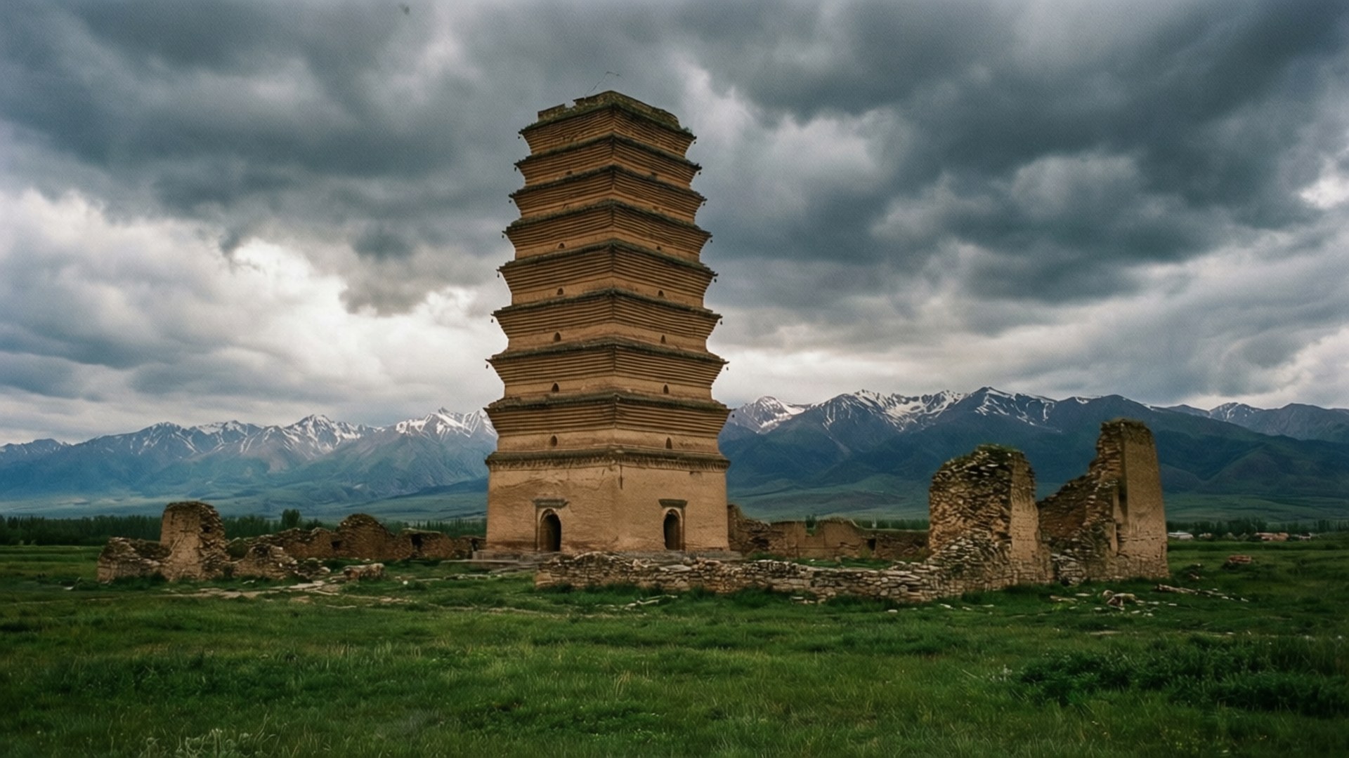 Burana Tower Ruins 壁紙 — 一座風化的古塔在長滿草的廢墟中高聳，遠處是白雪覆蓋的山脈，天空戲劇性地多雲。