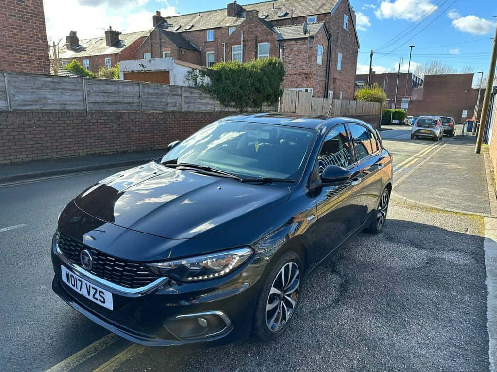 Fiat Tipo 桌布 壁紙 — 一台時尚黑色Fiat Punto停放在陽光明媚的住宅街道上，背景有磚造房屋和木柵欄。