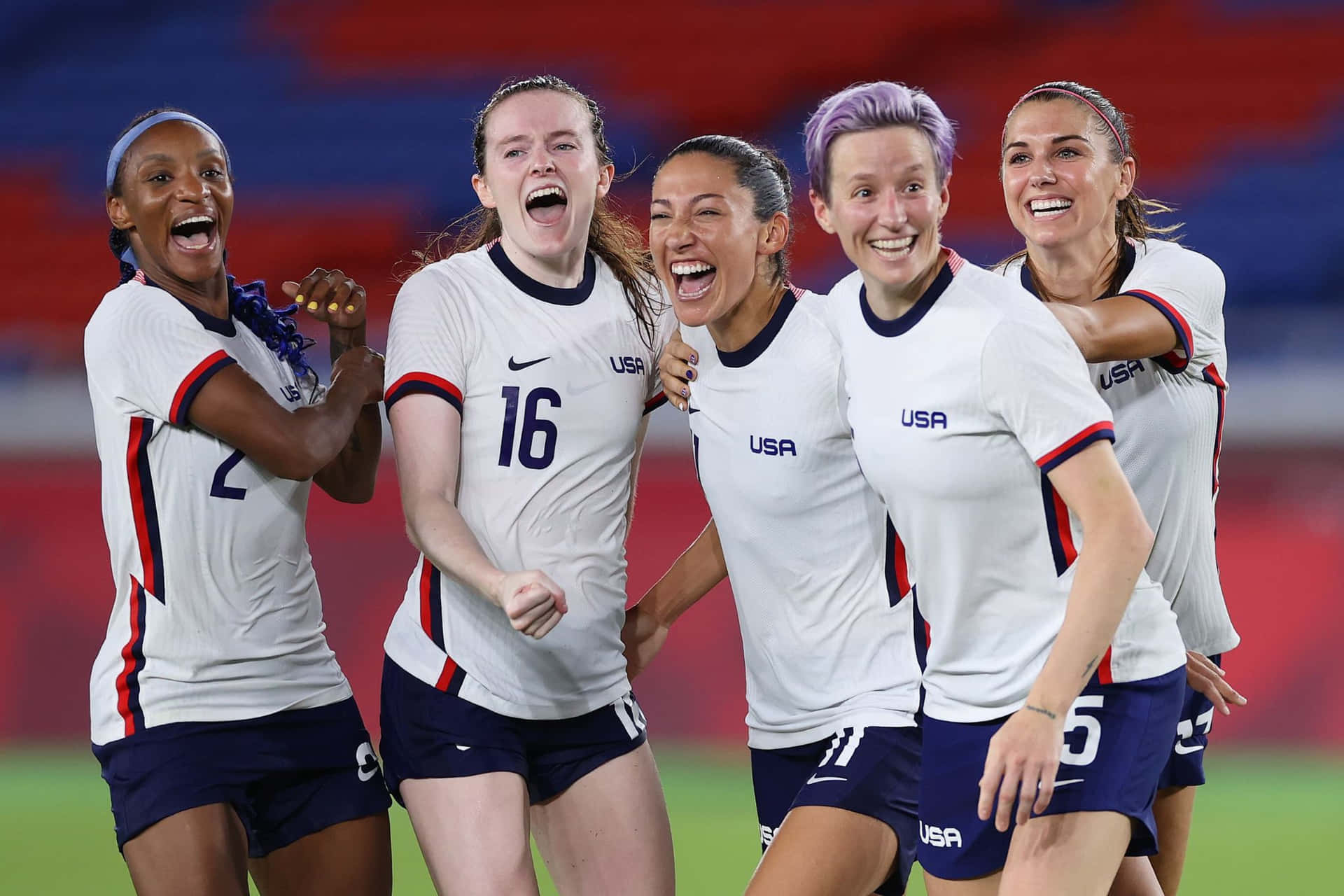  壁紙 — 五名身穿白色球衣、歡欣鼓舞的美國女子足球運動員在體育場上以燦爛的笑容和舉起的拳頭慶祝進球。