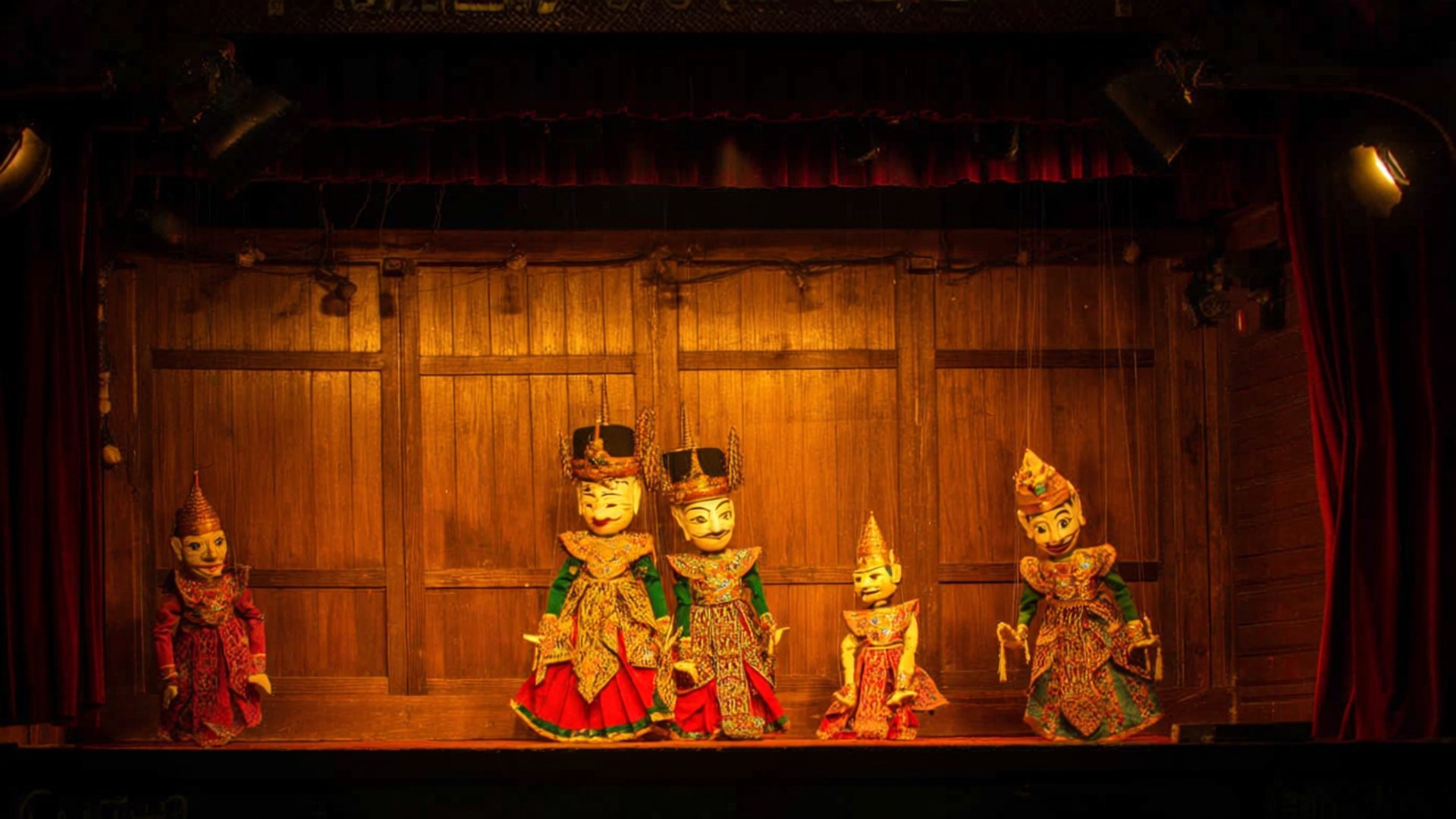 Burmese Puppet Show 壁紙 — 五個華麗的傳統木偶穿著鮮豔的服裝，在溫暖的劇院燈光下站在木製舞台上。