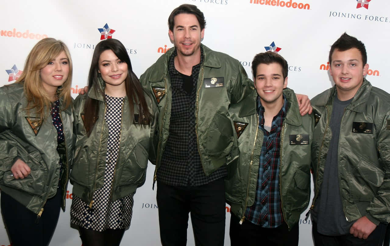  壁紙 — 五個微笑的人穿著相配的橄欖綠色外套，在一個 Nickelodeon 活動中，在有品牌標誌的背景前一起擺姿勢。