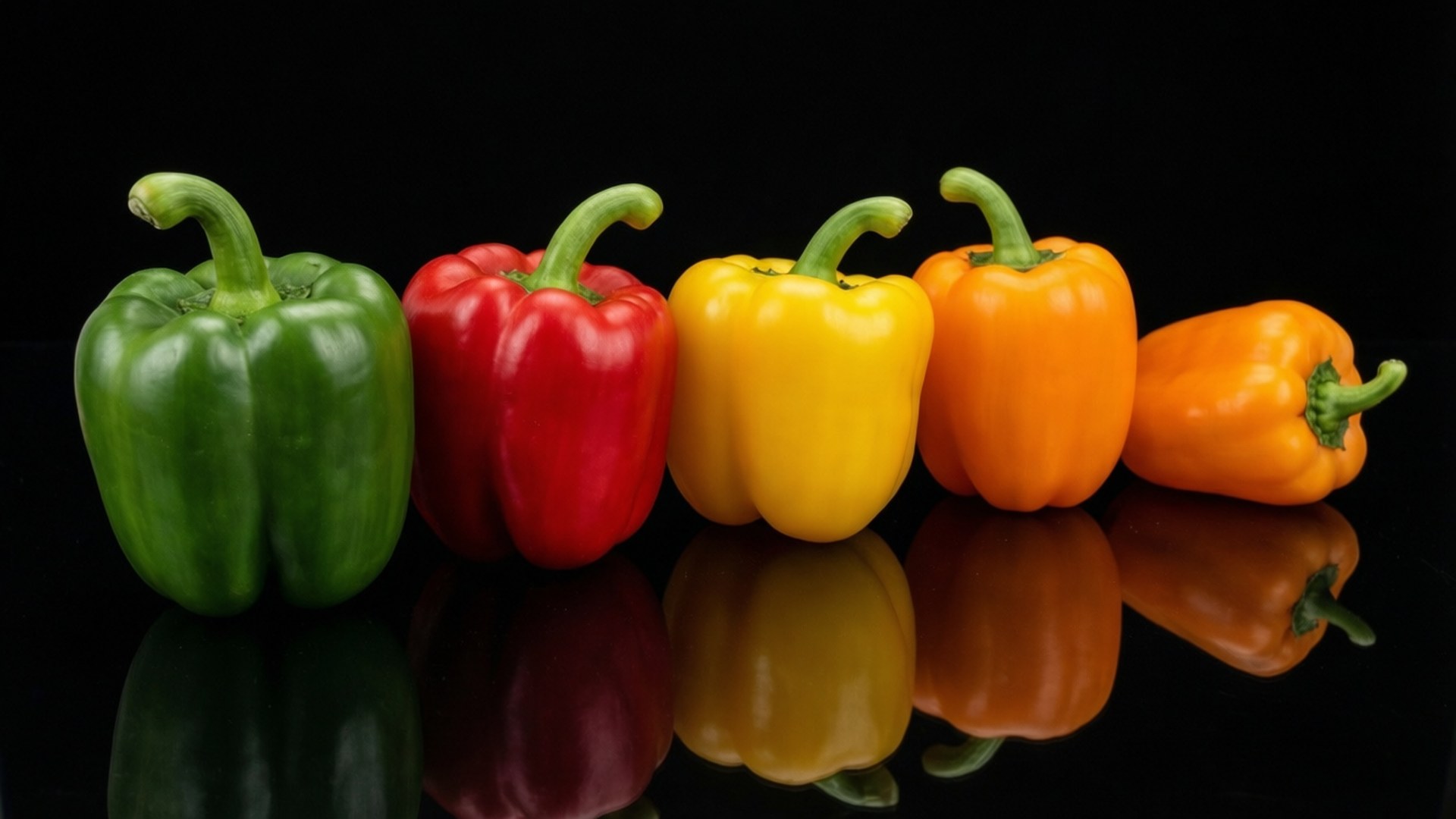 Pepper Lineup on Black 壁紙 — 五個鮮豔的燈籠椒，分別是綠色、紅色、黃色和橙色，排列在一排反光的黑色表面上。