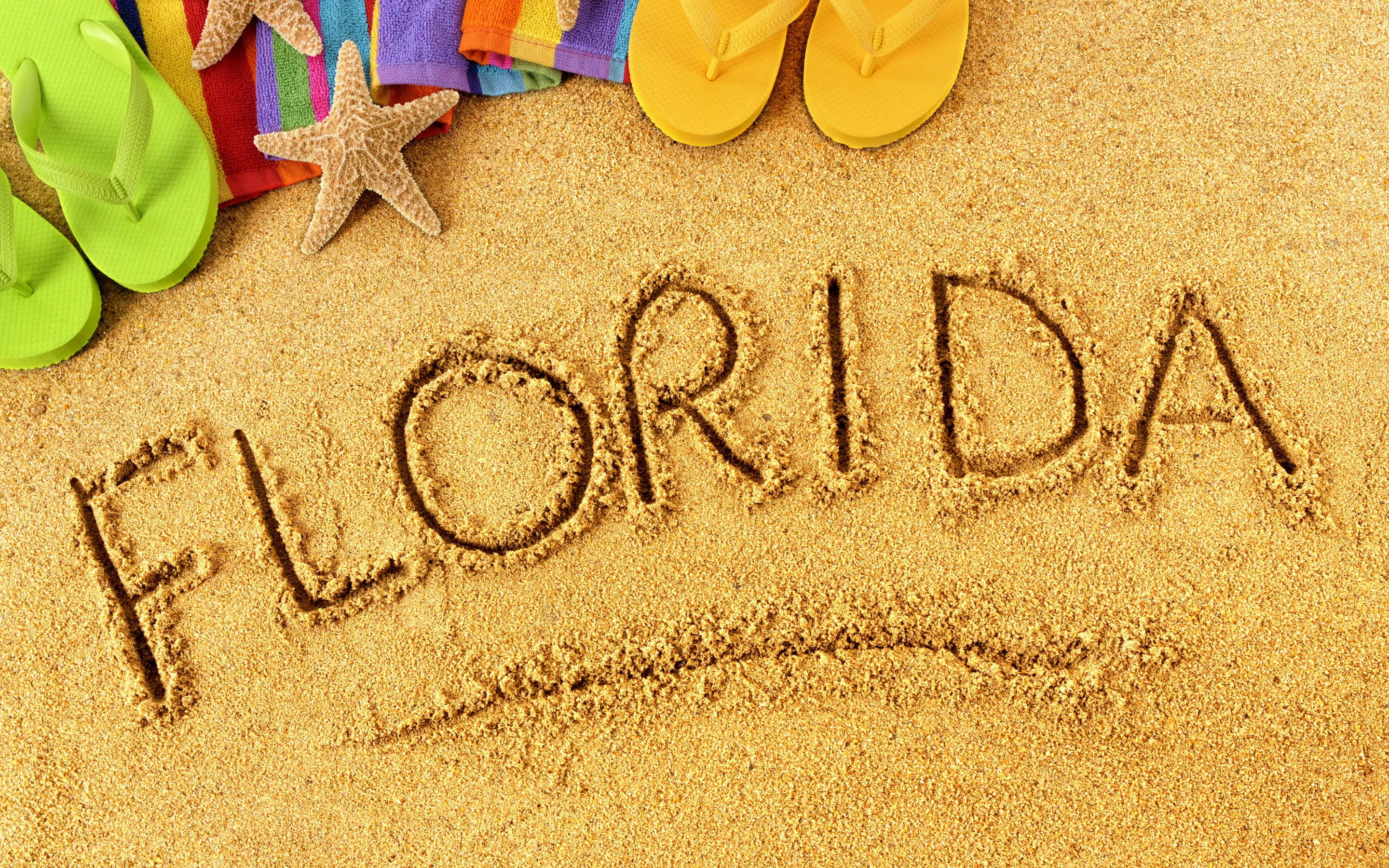 Beach Accessories 桌布 壁紙 — 「FLORIDA」這個字用金色沙寫在彩色夾腳拖、海星和彩虹毛巾旁邊。