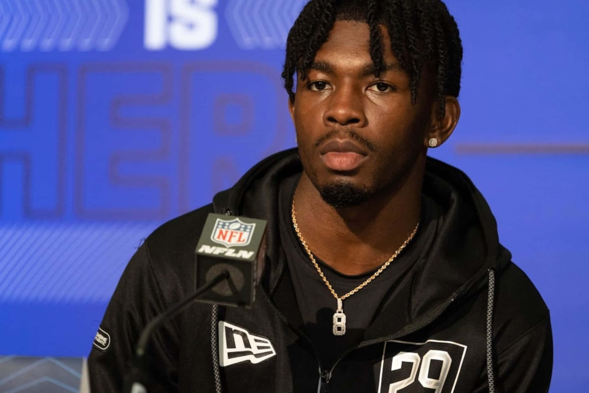 壁紙 — 一名專注的NFL球員身穿黑色連帽衫，號碼29，坐在記者會麥克風前，背景為藍色品牌背景。