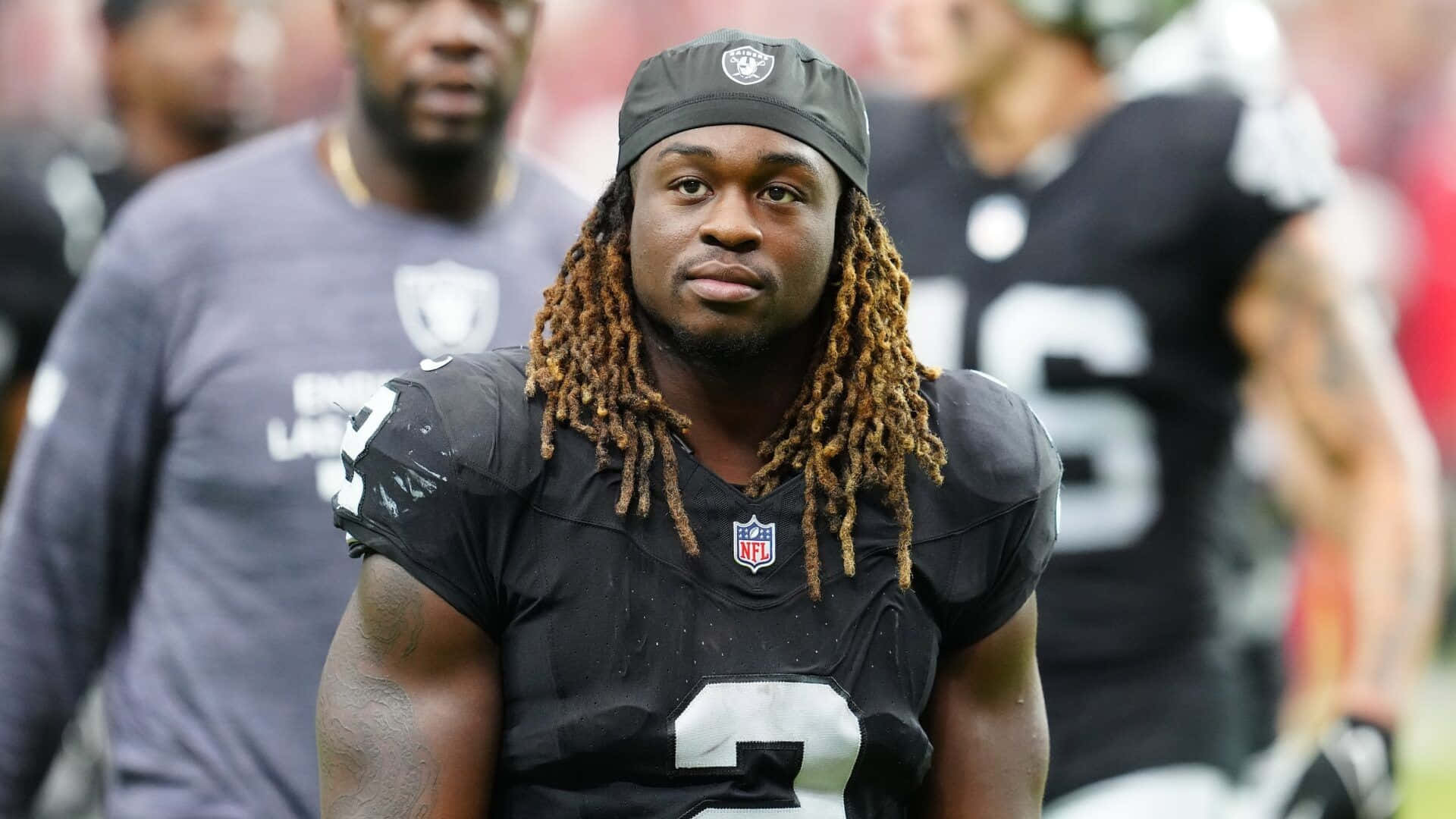 壁紙 — 一位專注的NFL球員，留著長辮子，在日間比賽中穿著黑色Raiders球衣凝視前方。