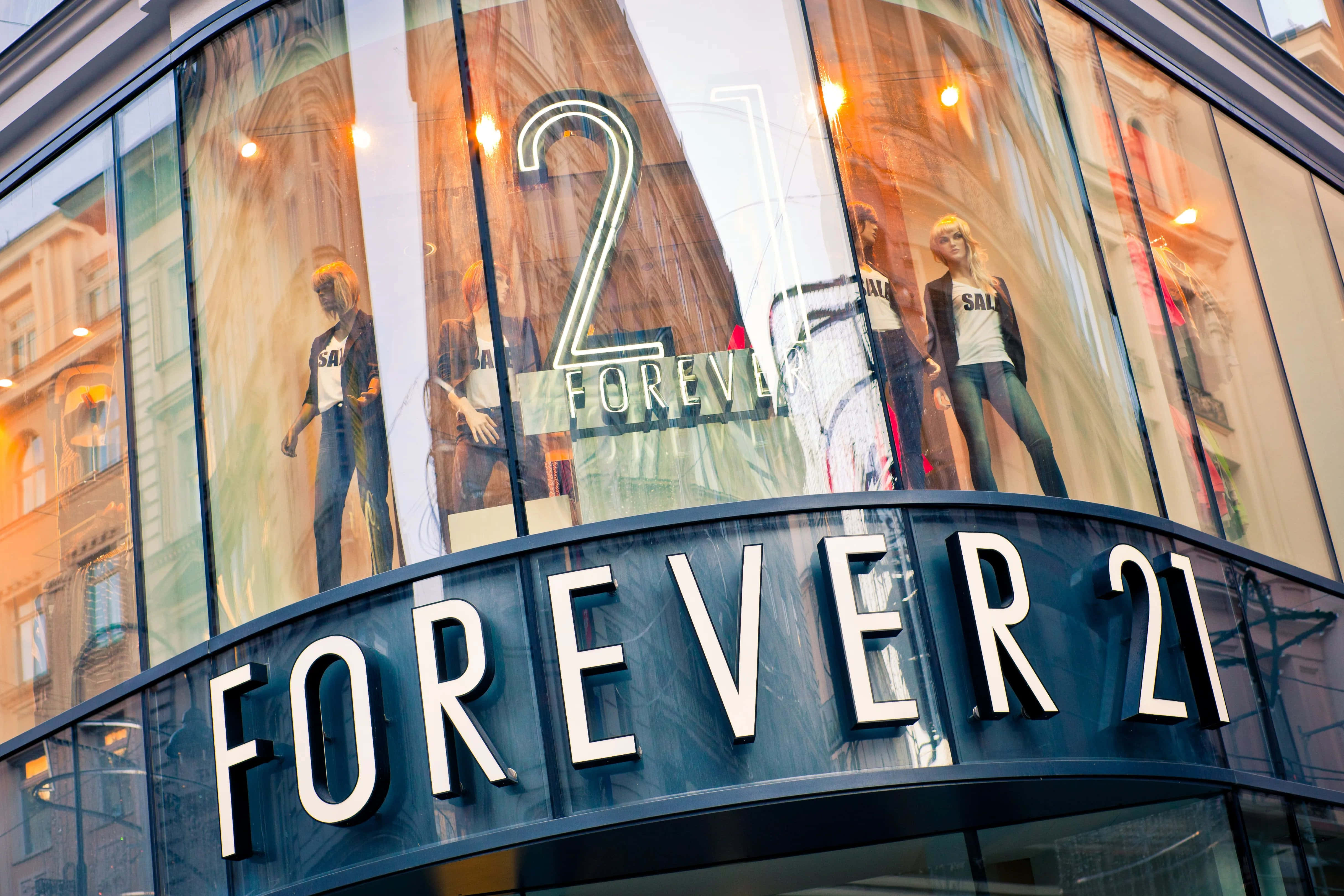 永遠21 桌布 壁紙 — Forever 21 的弧形玻璃店面展示穿著時尚服裝的人體模型，在溫暖的室內燈光下。