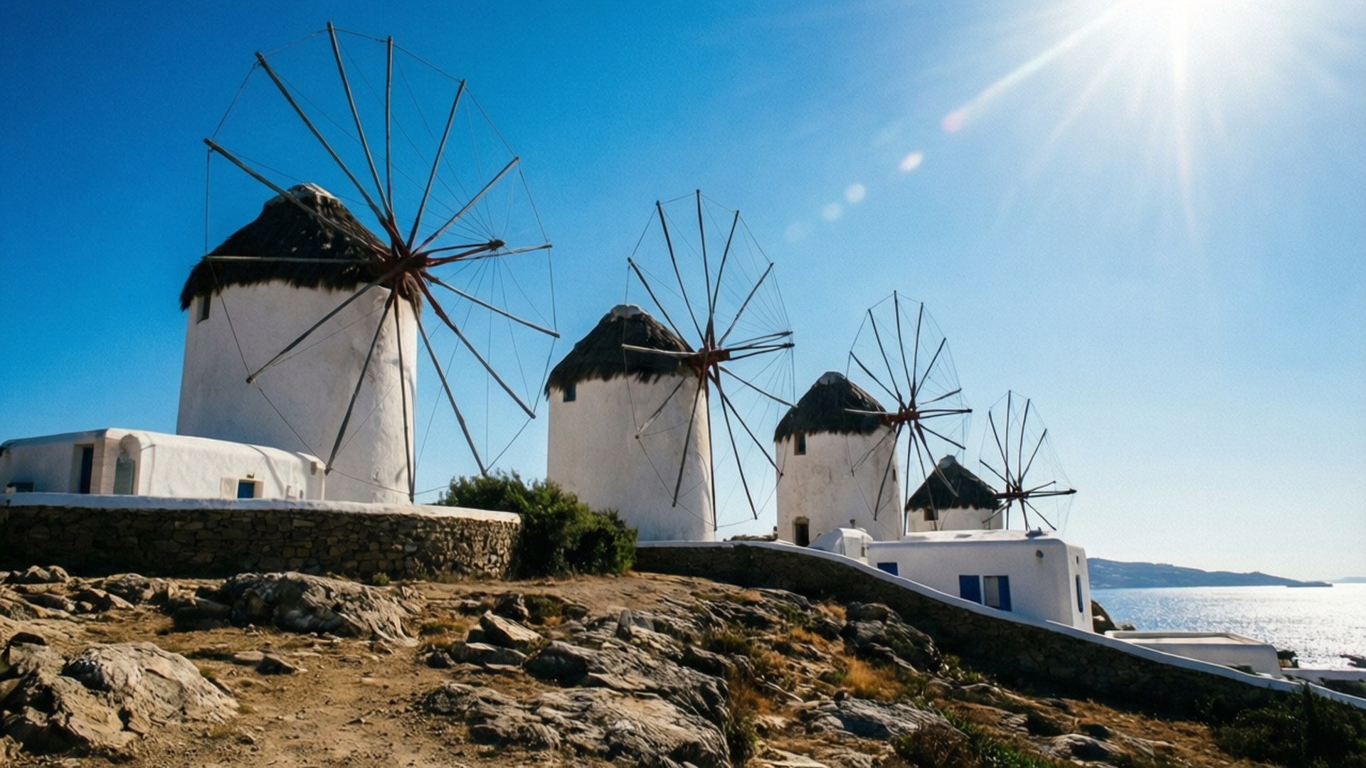 Mykonos Windmill Serenity 壁紙 — 四個標誌性的白色風車，帶有黑色茅草屋頂，矗立在陽光明媚的岩石山頂上，俯瞰著蔚藍天空下的大海。