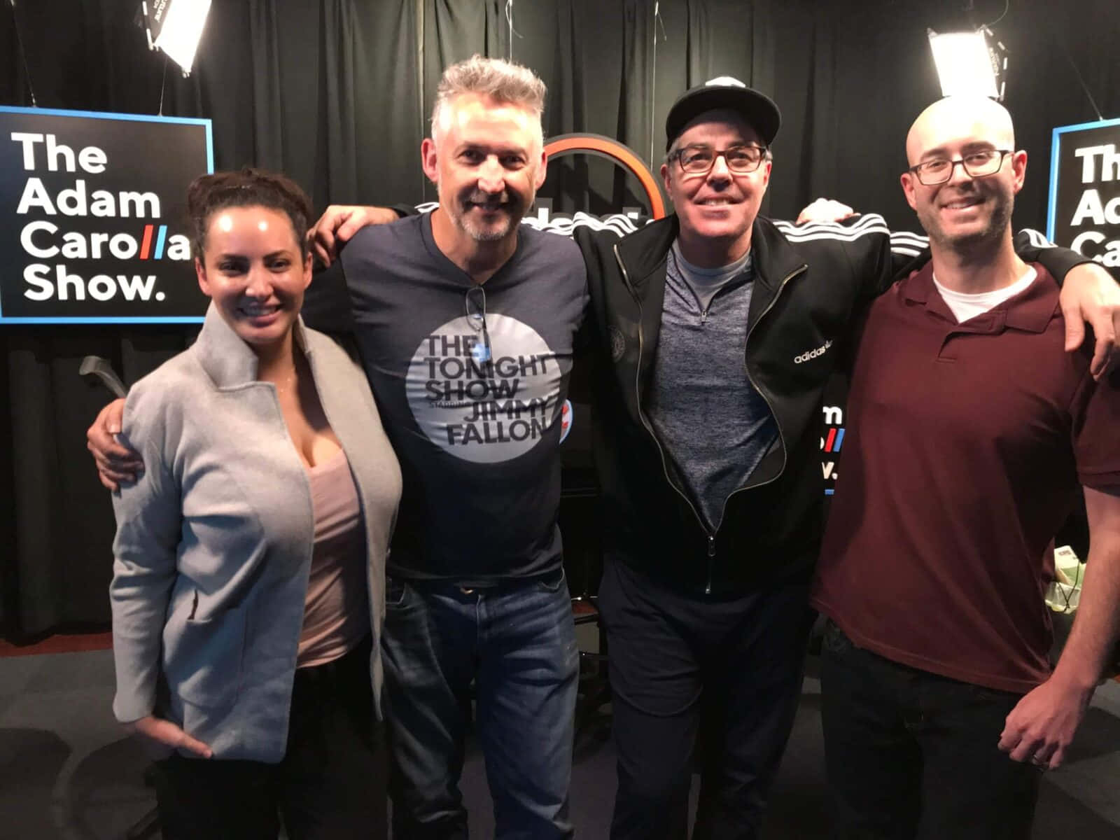  壁紙 — 四個微笑的人在有「The Adam Carolla Show」背景和明亮天花板燈光的攝影棚中一起擺姿勢。