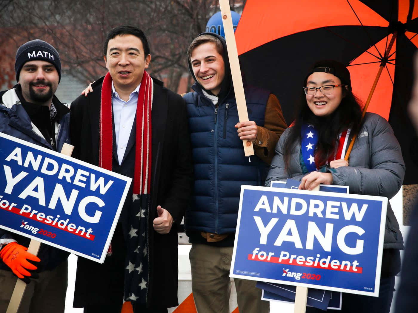 Andrew Yang 桌布 壁紙 — 四位微笑的支持者穿著冬季裝備，在橙色雨傘下戶外舉著藍色的Andrew Yang競選總統標語牌。