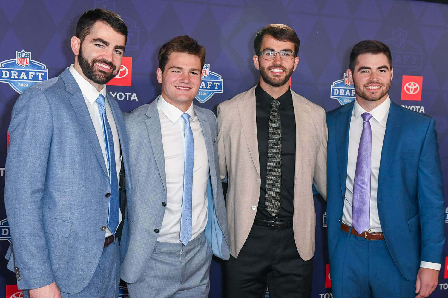  壁紙 — 四位身穿西裝的年輕男子在NFL選秀活動背景前自信地擺姿勢微笑。